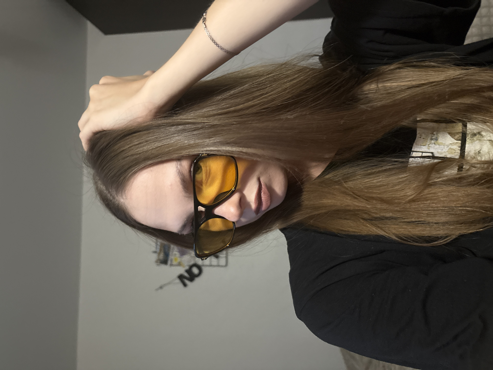 A person with long, light brown hair is shown from the chest up. They are wearing black clothing and yellow-tinted glasses. Their left arm is raised, with their hand resting on their head and touching their hair. A silver bracelet is visible on their left wrist. The background appears to be an indoor setting with light grey walls. In the background, there are some indistinct items on the wall, including what looks like a metal sign with the letters "NO" and possibly some other decorative elements. The lighting suggests it might be evening or indoors with artificial light. There are no strong indicators of the specific location within Perm, Russia, beyond it being an indoor environment. The person's expression is neutral.