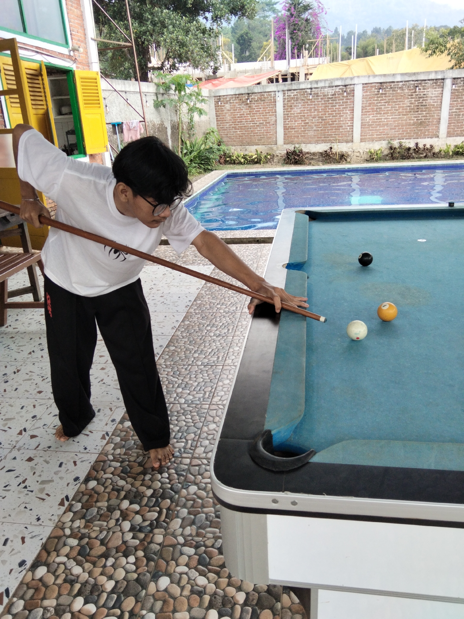 A young person with dark hair and glasses is playing billiards outdoors next to a swimming pool in Cisarua, Indonesia. The person is wearing a white t-shirt and black pants and is holding a pool cue, aiming at a white billiard ball. There are three other billiard balls on the table: a black ball and an orange ball. The pool table is blue and appears to be set up on a tiled patio area. In the background, a swimming pool with clear blue water is visible, along with a brick wall and some greenery. Construction can be seen in the distance, along with a yellow tarp. The overall scene suggests a leisure activity taking place in a tropical environment.