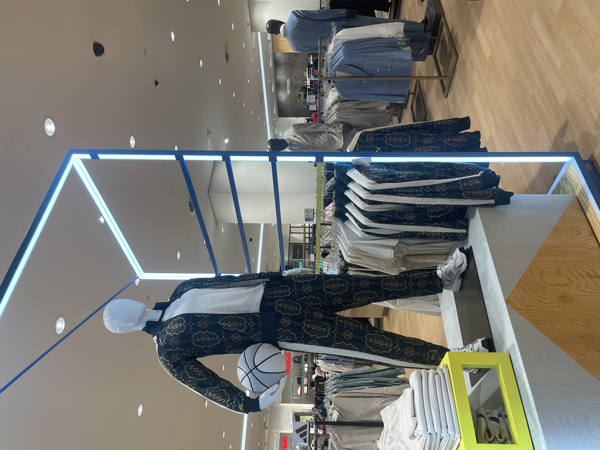 The photograph captures an interior scene, likely inside a clothing store located in Sofia, Bulgaria. In the foreground, a mannequin is dressed in a patterned suit, holding a basketball. Racks of clothing surround the mannequin. In the background, more clothing displays and mannequins are present. The lighting consists of both overhead recessed lights and linear LED fixtures. The overall ambiance suggests a modern retail environment.