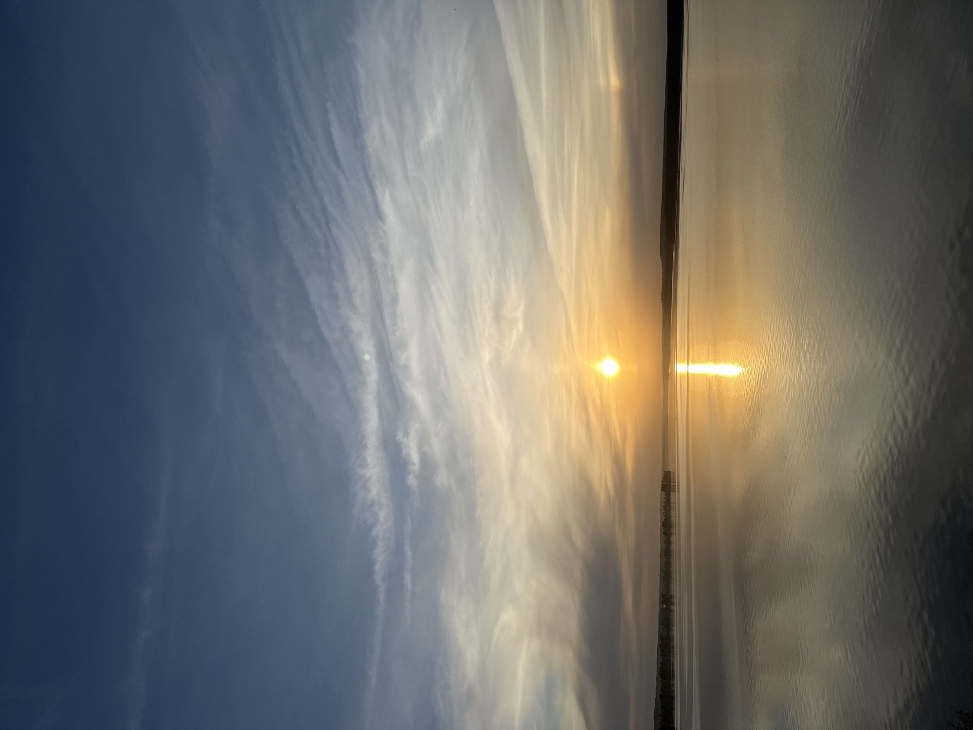 The image presents a sunset over water, with a clear sky grading into clouds. The sun is positioned at the horizon, emitting a bright light reflected on the water surface. The right side of the image is dominated by water. The left side shows a gradiated sky. A shoreline is visible in the middle of the image.
Due to the lack of visual indicators, there is no way to determine its location in the city of Votkinsk, Russia, with certainty.
