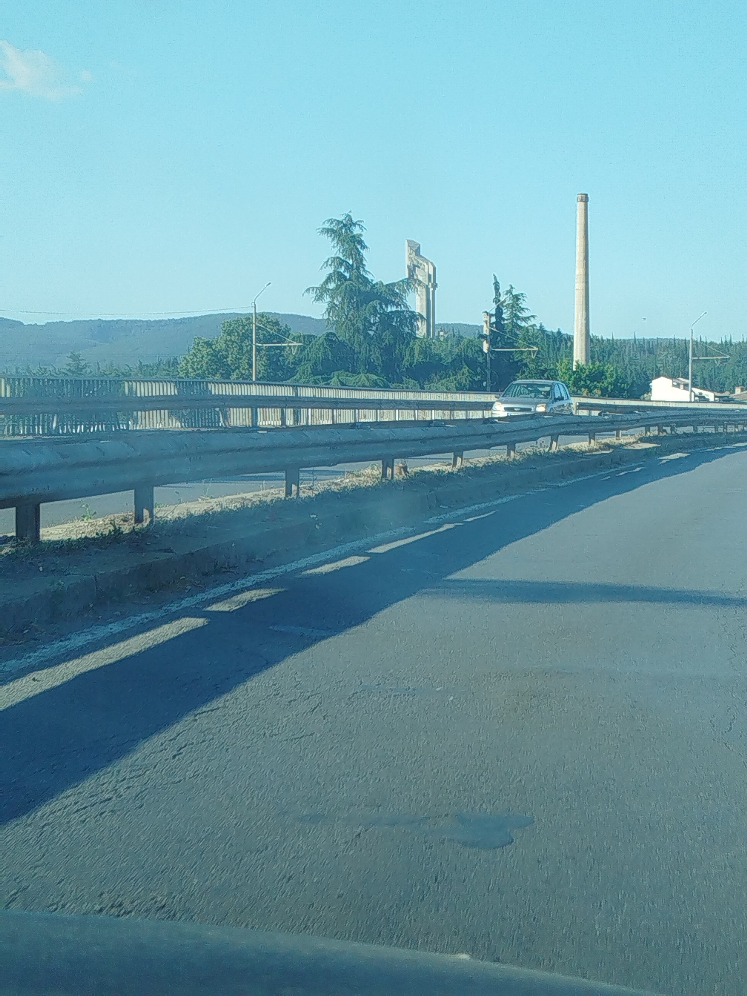 This is a landscape photograph taken from a moving vehicle on a sunny day. The scene depicts a highway overpass with a metal guardrail in the foreground. Beyond the overpass, a tall, cylindrical smokestack dominates the skyline, flanked by two imposing concrete structures that resemble monuments or industrial remnants. Lush green trees and hills form the backdrop, with a clear blue sky above.
A silver car is visible on the overpass, suggesting ongoing traffic. The sun is positioned to the right, casting long shadows across the road. The lighting indicates it is likely late afternoon or early evening. The environment appears to be semi-rural or industrial, given the presence of the smokestack and monumental structures. The overall mood is one of travel and observation, capturing a fleeting glimpse of the Bulgarian landscape. There is no visible text in the image.