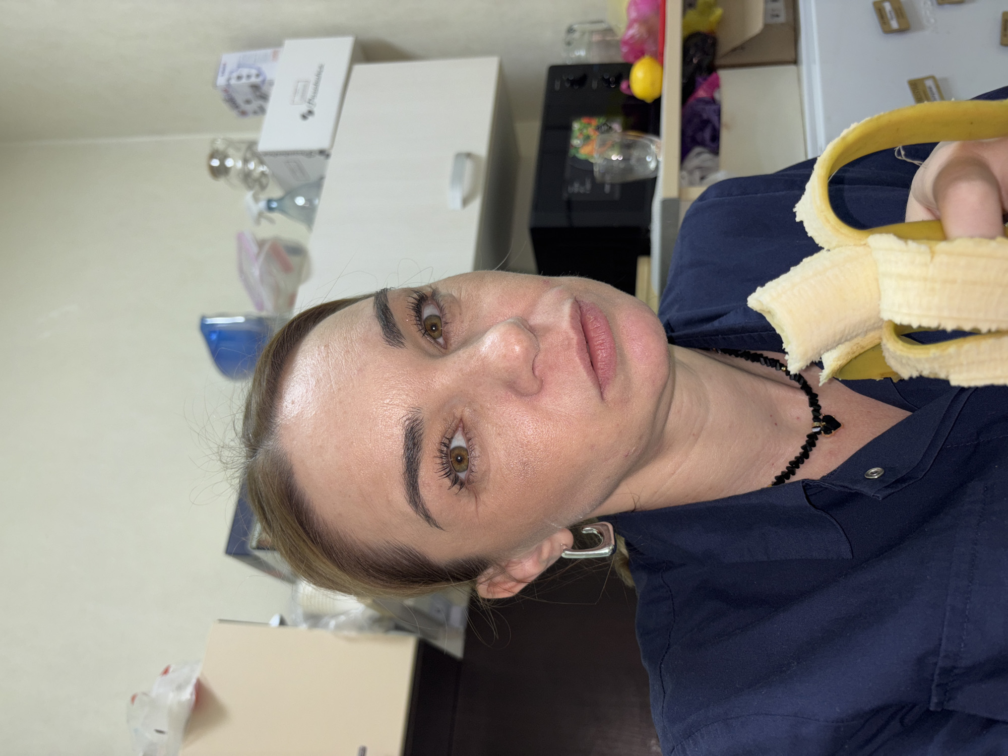 The image shows a woman indoors, likely in a kitchen setting. She is holding a partially peeled banana. The background features kitchen cabinets, a water dispenser, and other miscellaneous items. The city of Murino, Russia is not explicitly identifiable from the image.