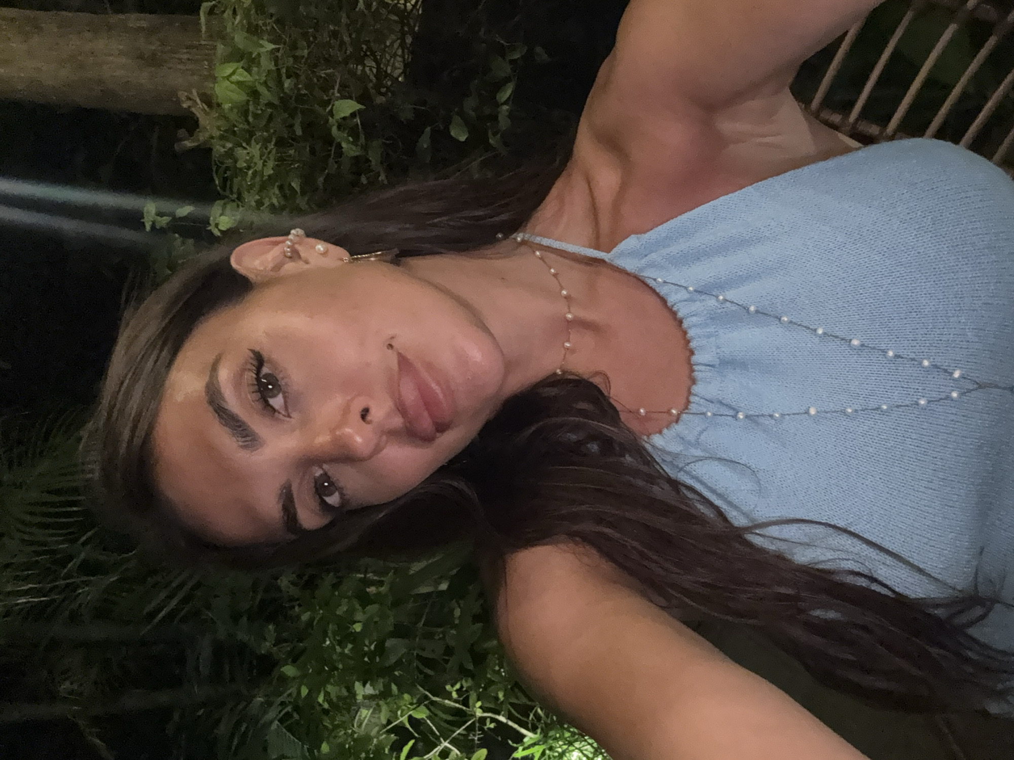 A young woman is posing in a selfie in Kuta Selatan, Indonesia. She has long brown hair and is wearing a light blue top with a pearl necklace. She is sticking her tongue out slightly. The background shows green foliage, possibly from a garden or park. The lighting suggests it is evening or night.