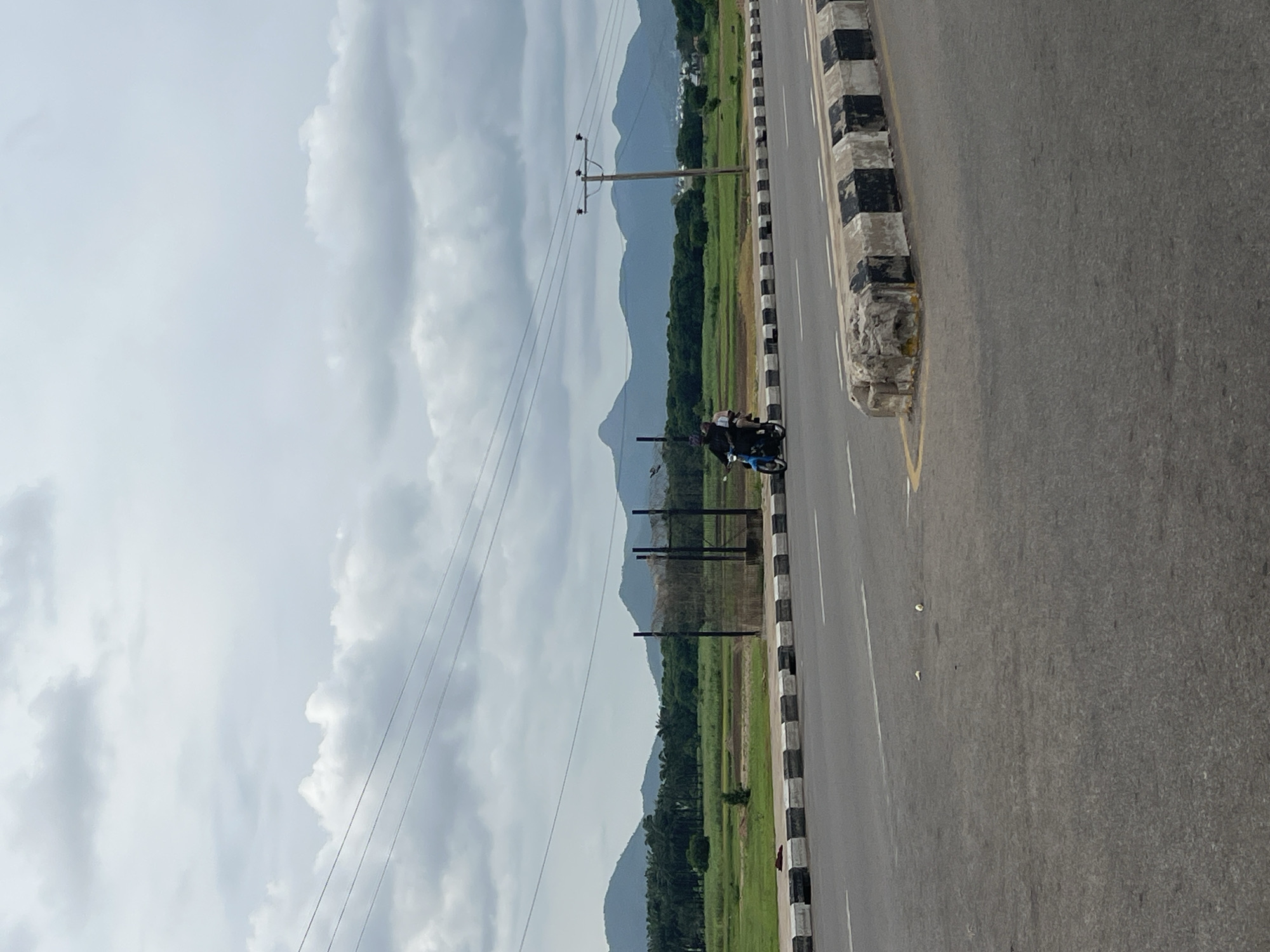 The image depicts a roadway scene with a cloudy sky overhead. In the foreground, a gray asphalt road is visible with white lane markings. A concrete divider with alternating black and white markings runs along the side of the road. In the middle ground, a blue motorcycle carries two individuals, one driving and the other as a passenger, moving away from the viewer. Beyond the road, a fence is situated, and further back, a vast green field stretches towards a line of trees. In the background, rolling hills and mountains under a cloudy sky form the horizon. The location is identified as Jalingo, Nigeria.