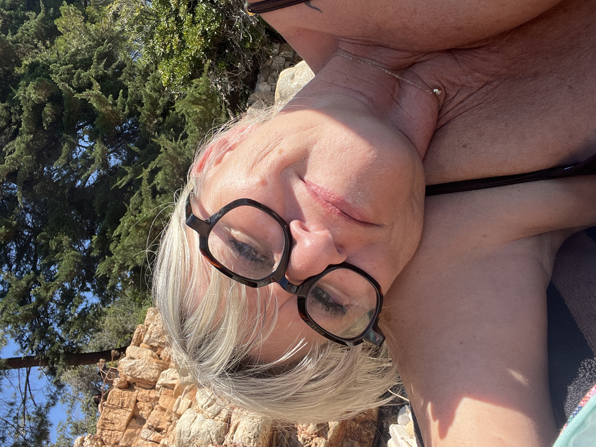 This is a close-up, overhead, selfie-style photograph of a woman with blonde hair and glasses. She is lying down outdoors, with a backdrop of trees, rocks, and a blue sky, suggesting a sunny day. The lighting indicates it is daytime. The woman is wearing a dark-colored bikini top. Her eyes are partially closed, and her mouth is slightly open, giving the impression of relaxation or contentment. A delicate silver chain with a small pendant is visible around her neck. The environment appears natural and possibly Mediterranean, consistent with the Cannes, France location context. There is no visible text in the image.