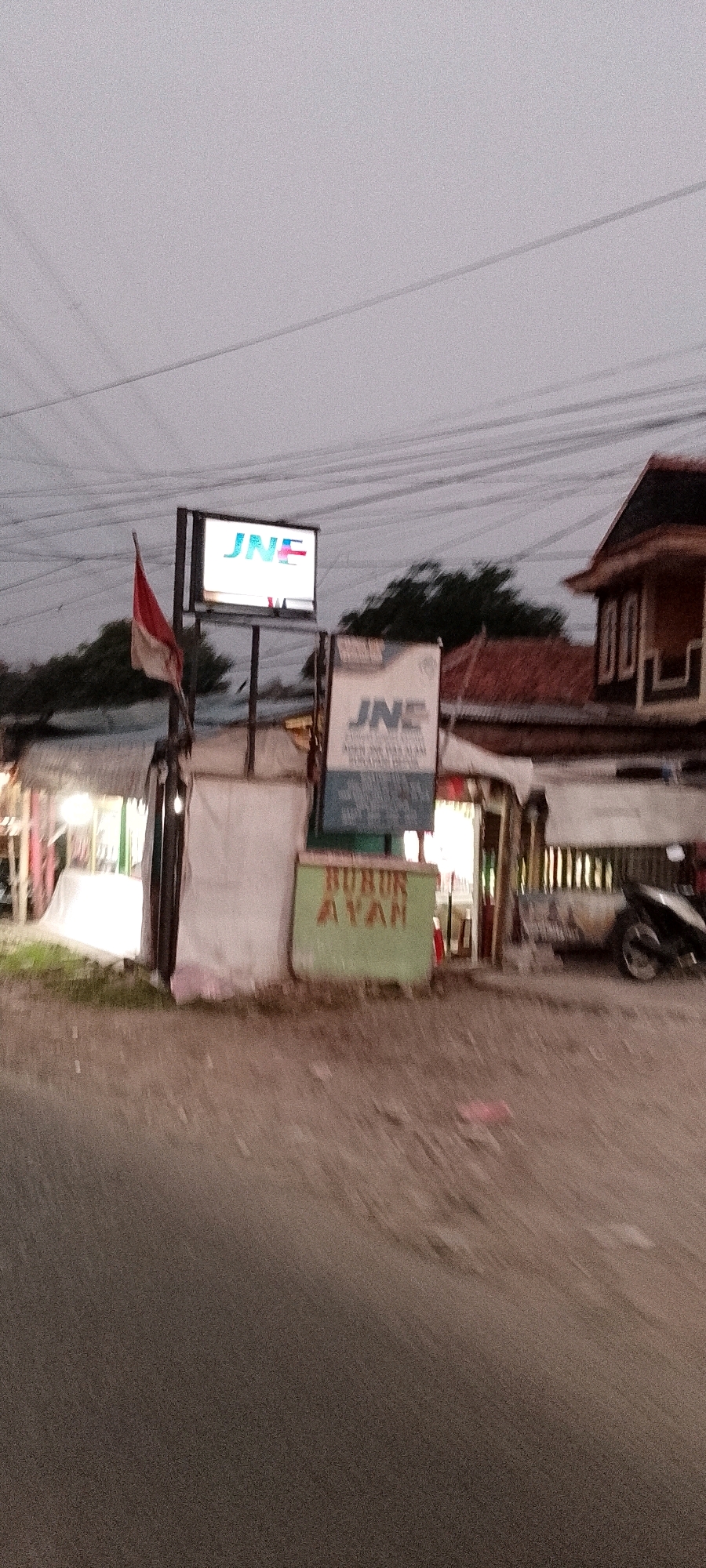 The image depicts an exterior view of a small commercial establishment in Depok, Indonesia, during what appears to be twilight. The scene is dominated by a bright, rectangular sign for "JNE," a logistics company, which is illuminated and positioned above another, larger sign that also features the "JNE" logo and what seems to be text in Indonesian. To the left of the JNE signs, a red and white Indonesian flag is affixed to a pole.
In front of the JNE signs, a green stand with red lettering that reads "BURUK AYAM" is visible. This likely indicates a food stall, possibly selling chicken porridge. The establishment itself appears to be a simple structure with a corrugated metal roof and white fabric covering some of the facade. To the right of the building, a motorcycle is parked. The ground in front of the establishment is a paved road, and overhead, power lines are strung across the sky, which is a muted grey color. The overall impression is that of a modest roadside business in an urban or semi-urban setting in Indonesia.