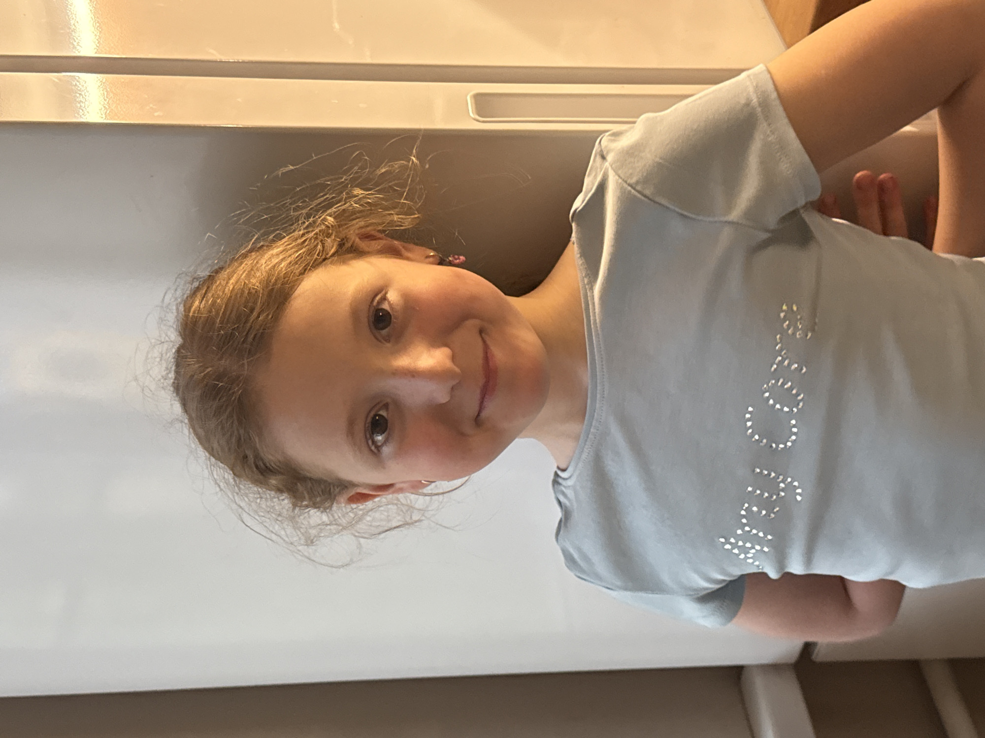 A young girl is posing for a portrait. She has light brown hair, fair skin, and a small smile. She is wearing a light blue t-shirt with glitter lettering. She is indoors. The background includes a white refrigerator and a white painted wall. The location is not identifiable as being in Rostov-on-Don, Russia, or anywhere else.
