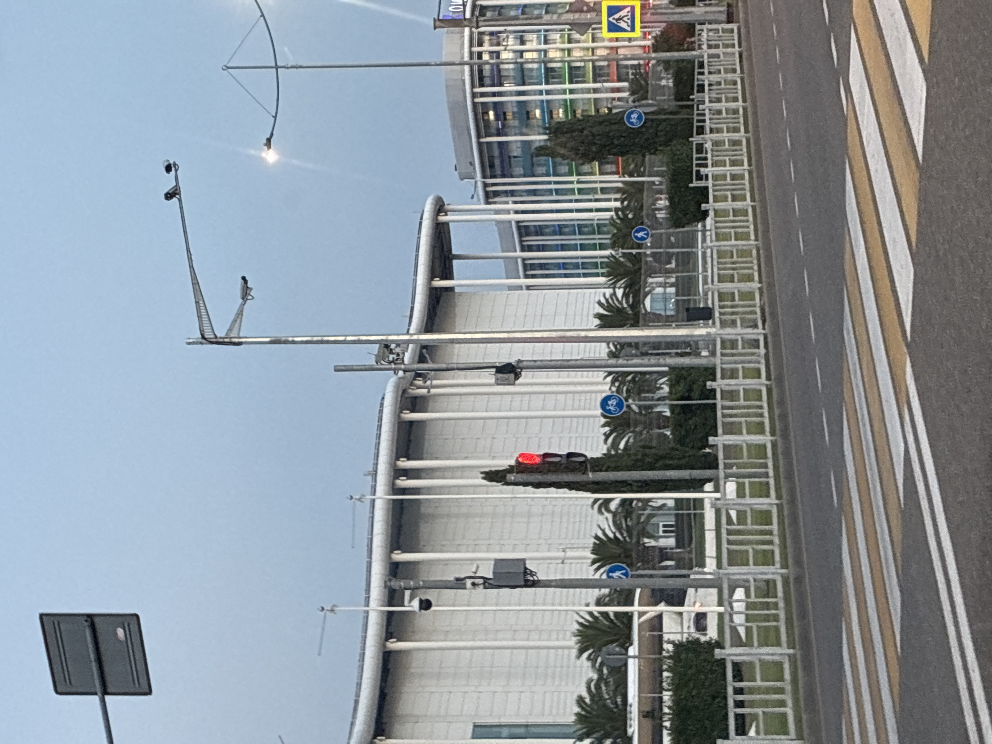 The image captures a street scene in Sirius, Russia, on a clear, overcast day. In the foreground, a pedestrian crossing with yellow and white stripes is visible on asphalt. Beyond the crossing, a white metal fence runs along the sidewalk, separating it from a green hedge.
Several traffic signals and signs are present. A traffic light displays a red signal. Two blue circular signs with white pictograms indicating a pedestrian crossing and a bicycle path are also visible. To the right, a yellow square sign with a black pedestrian pictogram is mounted on a pole.
Several palm trees and cypress trees are planted along the sidewalk. In the background, a large, modern building with a white facade and numerous vertical white columns dominates the scene. To the right of this building, another multi-story building with a rounded facade and colorful windows is situated. Streetlights and security cameras are mounted on poles throughout the area. The sky is a pale blue, indicating either early morning or late evening. No people are clearly visible in the image.
