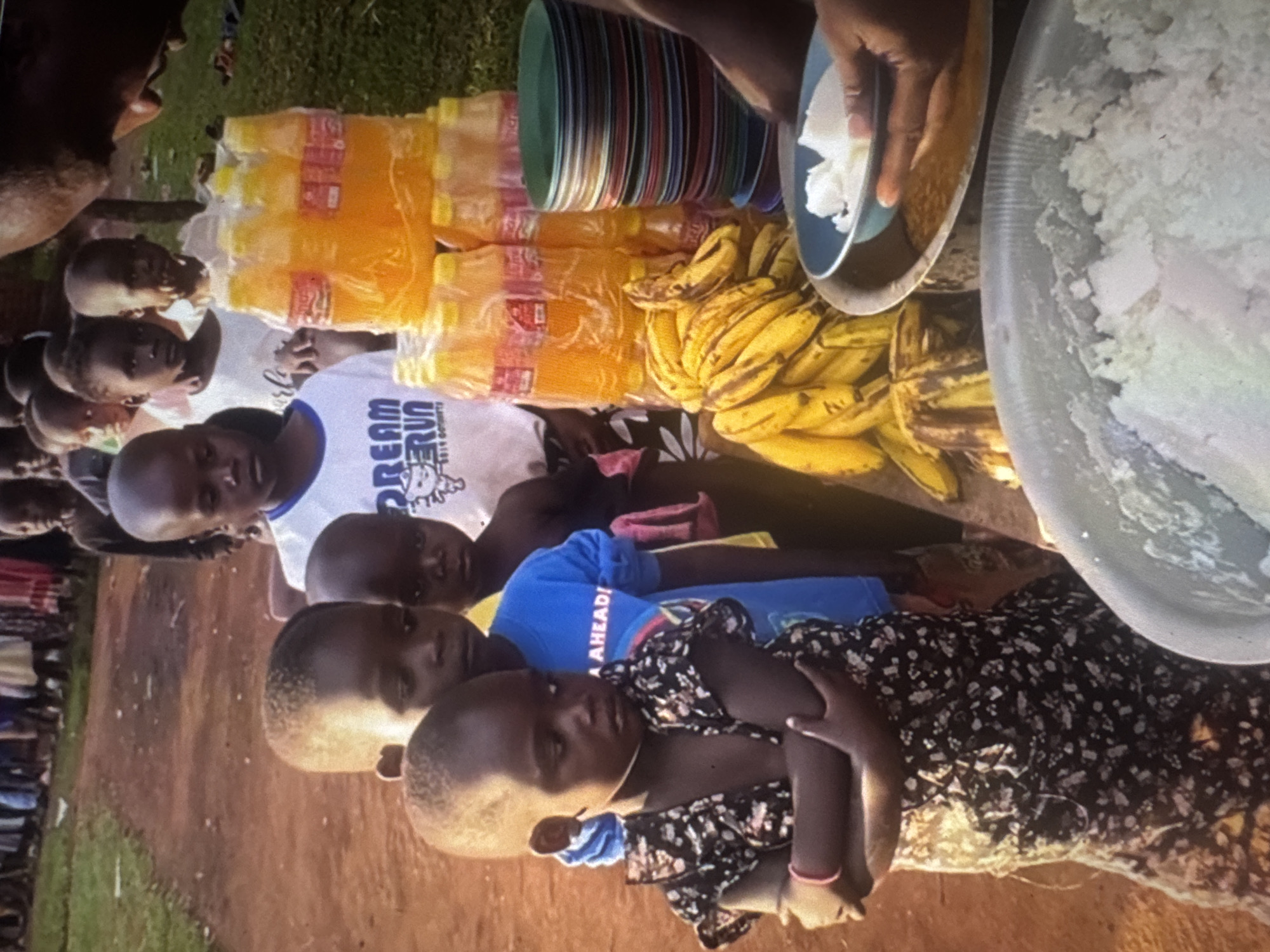 The image depicts a group of children and adults in what appears to be an outdoor gathering, possibly a food distribution event. The setting is dusty with a dirt path in the background and some green grass on the right. In the foreground, a young child in a dark floral dress stands with their arms crossed. Behind this child, other children are lined up, some wearing t-shirts with writing. Further back, a crowd of people, including adults and more children, are visible.
On a table or surface in the middle ground, there are stacks of plastic bottles containing an orange liquid, bunches of bananas, and a stack of colorful plates. A hand is seen holding a blue bowl filled with white material, possibly food, over a larger silver basin also containing a white, granular substance. The overall scene suggests a communal meal or aid distribution, possibly in a less developed urban or rural area. The presence of the dirt path, the simple serving setup, and the gathering of people imply a local community event. No specific landmarks or indicators place this scene in Abuja, Nigeria.
