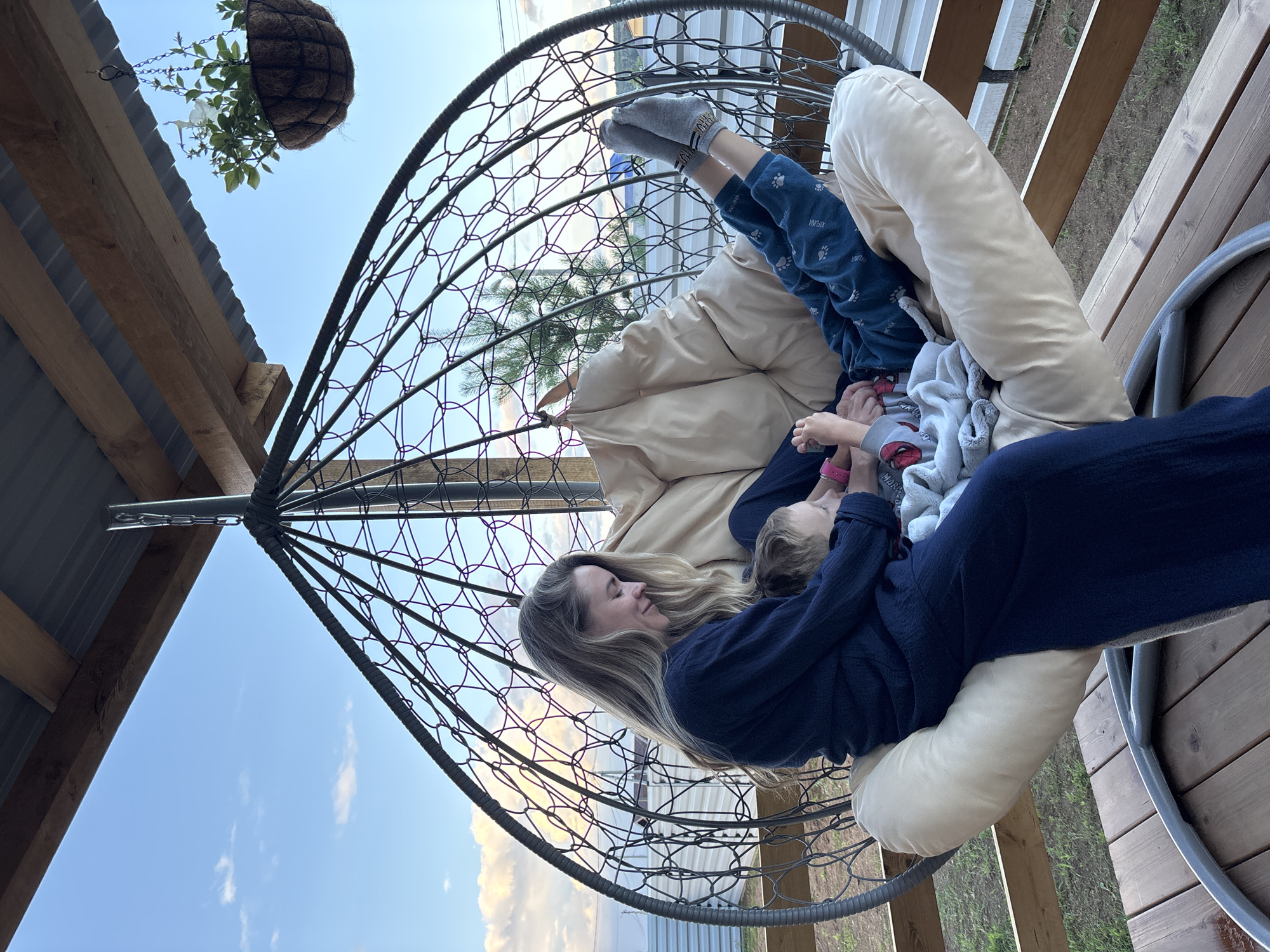 A woman and a child are relaxing in a hanging chair on a wooden deck. The woman, with long blonde hair, is wearing a navy blue top and is lying down, looking towards the child. The child, wearing blue pants with paw prints and grey socks, is lying on top of the woman and is looking at something in their hands. A beige pillow is supporting the child's legs. Behind them, a hanging basket with a plant and white flowers is visible against a clear blue sky with some clouds. The setting appears to be outdoors, possibly a backyard or patio, with wooden decking and structural elements of a roof overhead. The scene suggests a calm and peaceful moment between the two individuals, possibly late afternoon or early evening given the lighting. No visible text is present in the image.