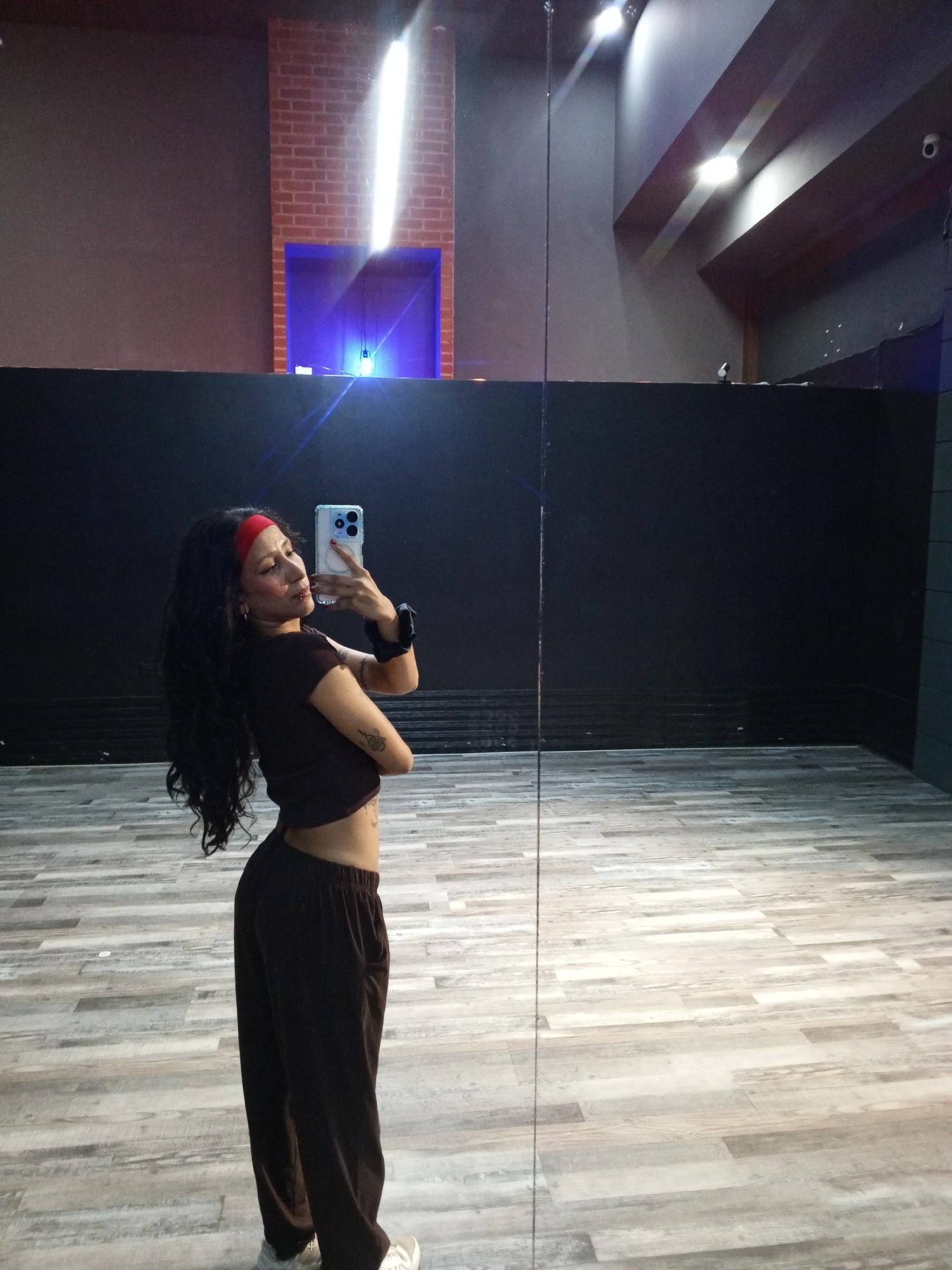 The image shows a young woman in a dance studio. She is standing at an angle to the camera, holding a smartphone up to take a selfie. She is wearing a brown crop top, loose brown pants, and a red headband. She has long, dark, wavy hair. Her left arm is visible, and she has a tattoo on her forearm. The background is a dance studio with a wood-look floor and a black wall. There is a brick structure with blue lighting on the wall. The lighting is dim, and there are spotlights on the ceiling.
The image does not provide sufficient information to determine the specific location in Pampatar, Venezuela.
