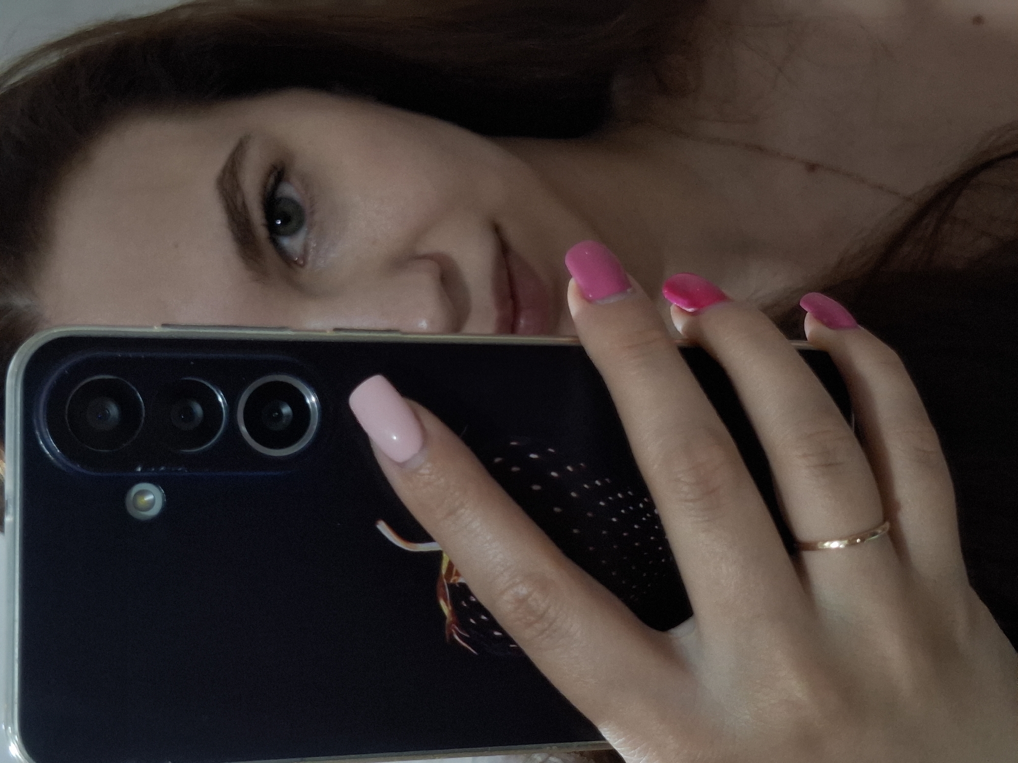 A woman with light skin, brown hair, and pink painted fingernails is holding a black smartphone with a clear case in her right hand. The phone partially obscures her face, but her left eye, nose, and lips are visible. The camera lenses and flash of the phone are visible in the upper left corner of the frame. A gold ring is on the fourth finger of her right hand. The background is blurry. There is no evidence that places the subject in the city of Chaikovsky, Russia.