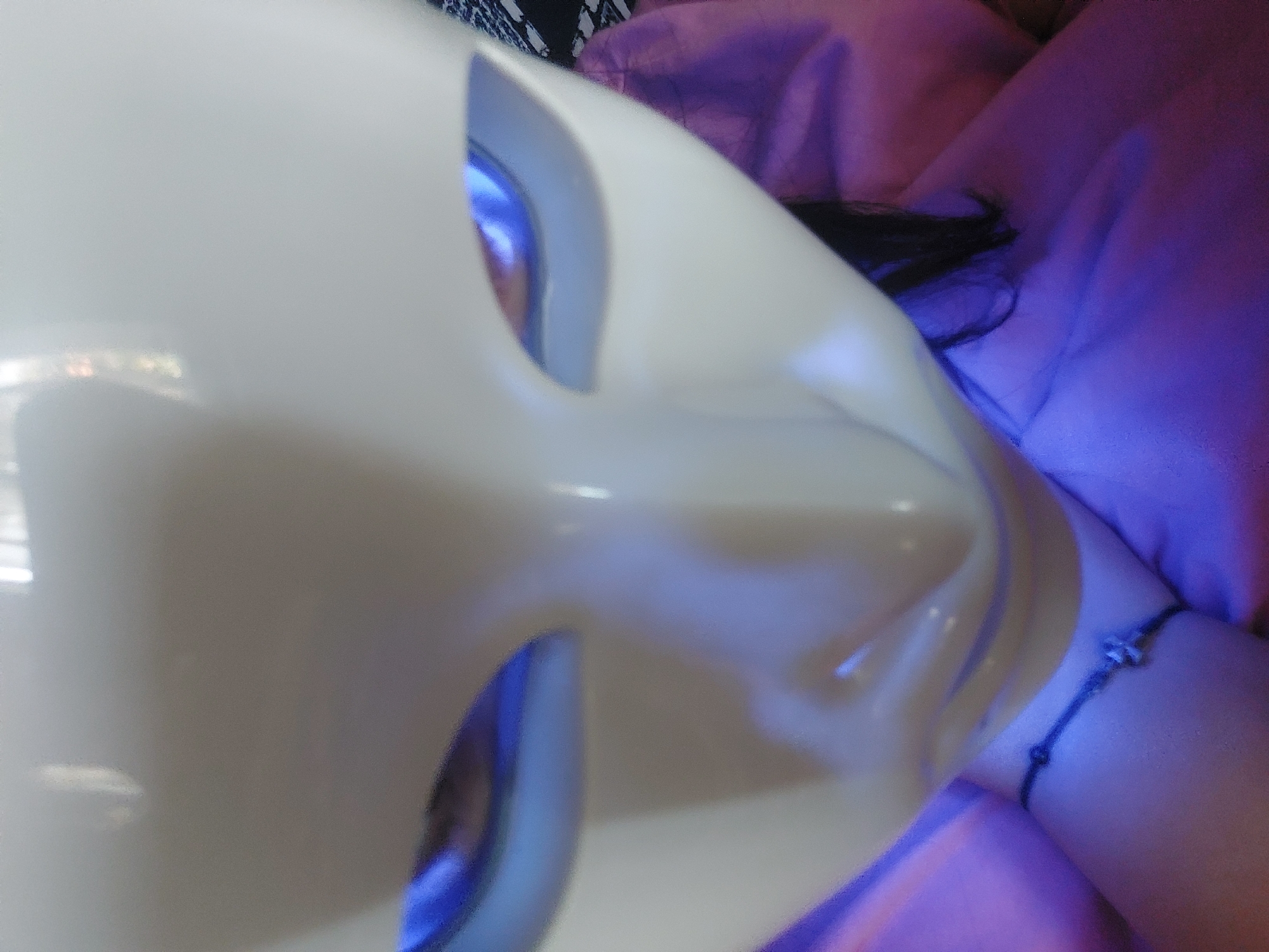 A close-up, high-angle view depicts an individual wearing a smooth, matte white mask that covers the upper portion of the face. The mask features elongated eye cutouts, behind which a strong blue light emanates, illuminating the wearer's eyes from within. The mask has a prominent, stylized nose ridge and subtle lip forms. Below the mask, the chin and neck of the wearer are visible. A bare forearm is positioned horizontally below the mask, adorned with a thin black string bracelet featuring a small, silver-toned, cross-shaped charm. Strands of dark hair are visible on the right side of the image, emerging from behind the mask. The person and surrounding environment are bathed in a magenta or deep purple light, which strongly tints the visible fabric, possibly clothing or a blanket, in the lower and right portions of the frame. A fragmented pattern of black and white fabric is partially visible behind the right side of the mask.