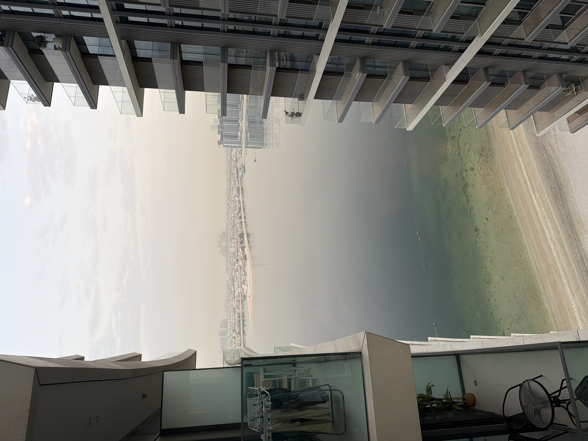 The image shows a view from a balcony of a modern apartment building in Dubai, United Arab Emirates. The balcony on the left features a clothes drying rack with clothes, a potted plant, and a small table with two chairs. The building on the right is a high-rise with multiple balconies. In the background, there is a body of water, a beach, and other buildings, suggesting an urban coastal setting. The sky is overcast, and there is a hazy atmosphere. The architecture and the urban landscape are characteristic of Dubai.