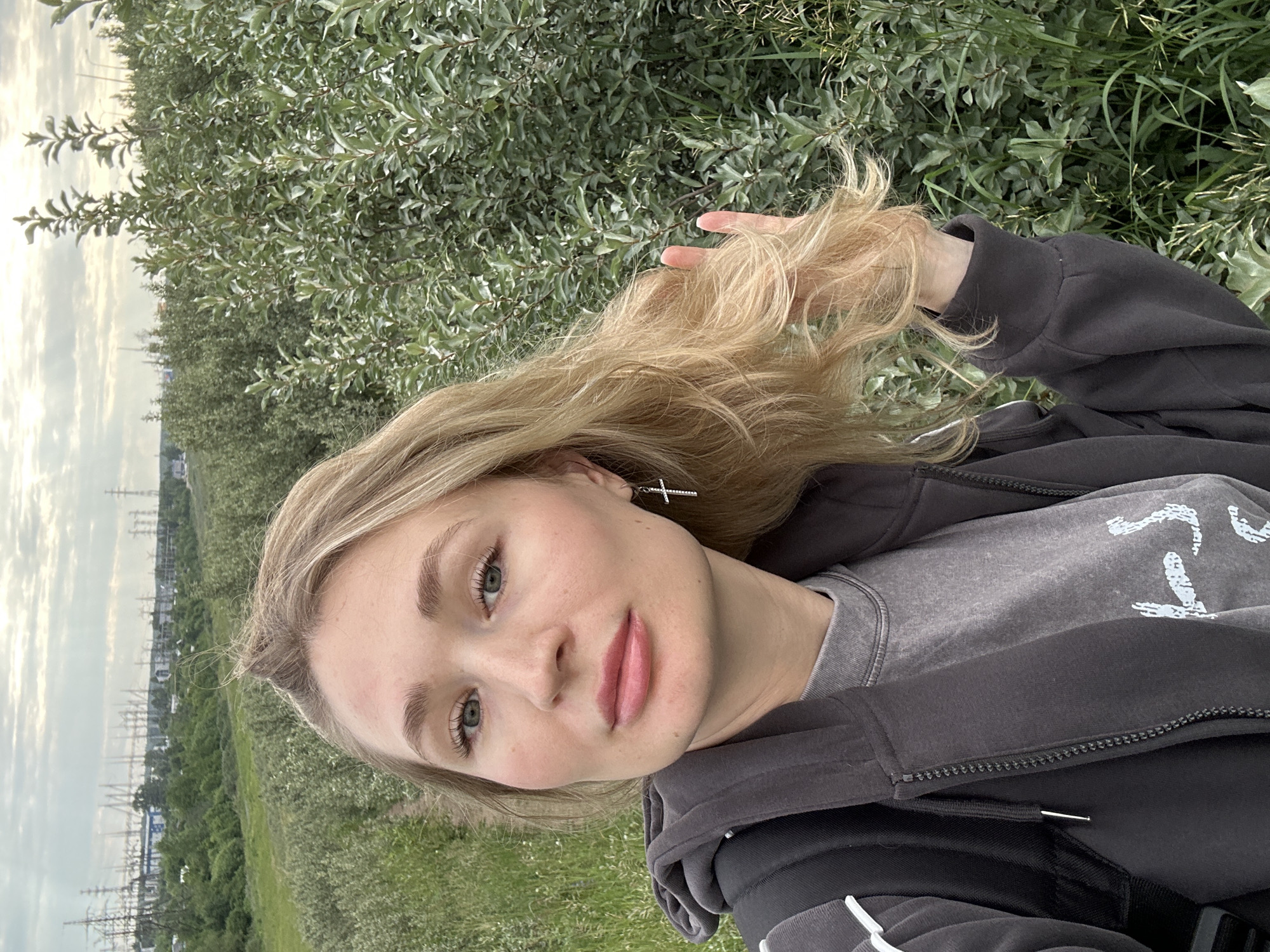 A young blonde woman is taking a selfie outdoors, with a backdrop of green foliage and distant industrial structures. The woman is wearing a dark grey hooded jacket over a grey t-shirt. She has a silver cross earring in her right ear and is touching her hair with her left hand, which is held up near her face. In the background, there is a vast expanse of green trees and bushes, and beyond that, a clear sky with some clouds. Further in the distance, the silhouette of electrical transmission towers and buildings can be seen, suggesting an industrial area on the outskirts of the city. The overall setting appears to be a natural area adjacent to an industrial zone, possibly on the outskirts of Chaikovsky, Russia, given the presence of such infrastructure.