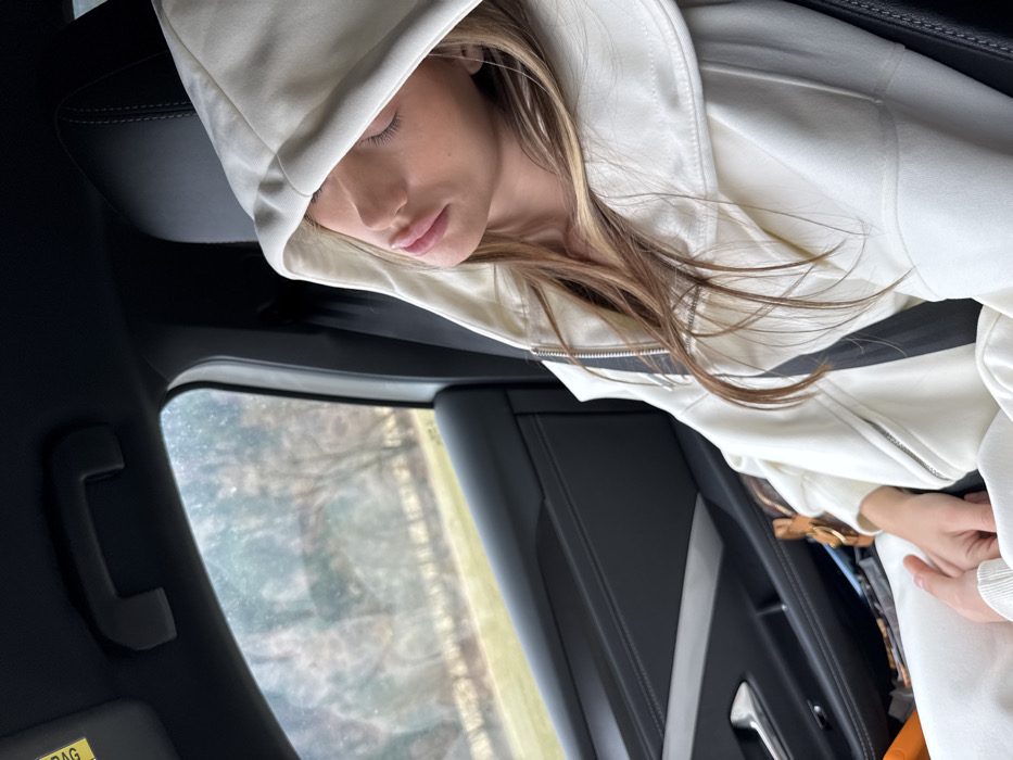 A person is depicted asleep in the front passenger seat of a modern vehicle, traveling through a rural or semi-rural area near Murau, Austria. The individual, appearing to be a young female, is wearing an off-white hooded sweatshirt with a visible zipper and matching sweatpants. Her hood is pulled up, obscuring the upper portion of her face, while her eyes are closed and lips slightly parted. Long, light brown hair with blonde highlights is visible around her face. A dark seatbelt is secured across her chest and lap. The vehicle's interior features dark gray or black upholstery on the seat and door panel. A dark gray grab handle is mounted above the side window, and a sun visor with a yellow "AIR BAG" warning is partially visible in the upper left. Through the blurred side window, bare deciduous trees and green ground are visible, suggesting a natural landscape.