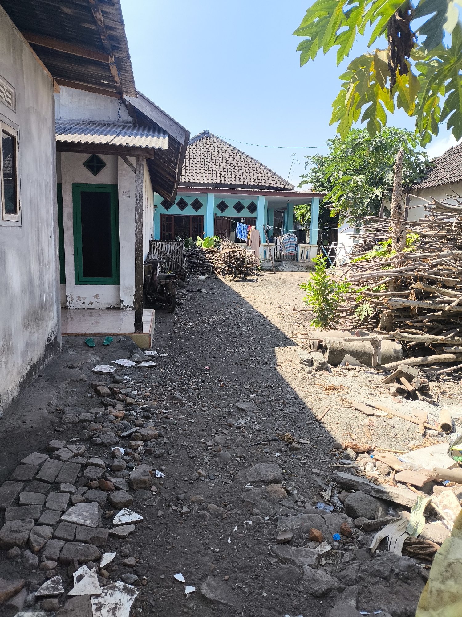 The image depicts an outdoor scene on a sunny day, likely in Grati, Indonesia, showing a somewhat unkempt yard with two houses and various scattered objects. The foreground is dominated by a dirt path covered with small stones, gravel, and broken pieces of concrete and tile. To the left, a grey-colored house with a white trim and a window with a light-colored frame is visible, its lower section showing signs of wear. Adjacent to it is another section of a building with a corrugated metal roof supported by wooden beams, and a green-framed doorway. To the right of this structure, a blue house with a patterned roof and teal diamond-shaped decorative elements on its walls is partially visible. Laundry is hanging on a clothesline in front of the blue house.
Further to the right, a large pile of dry branches and sticks is stacked against a wooden post and a rough fence. Several other miscellaneous objects, including a motorcycle, a wheelbarrow, concrete pipes, and more debris, are scattered around the yard. A small green plant grows near the pile of branches. In the background, more trees and vegetation are present, along with parts of other buildings. The sky is clear and blue. The overall impression is of a residential area with a focus on functional, perhaps less-than-pristine, outdoor living spaces.