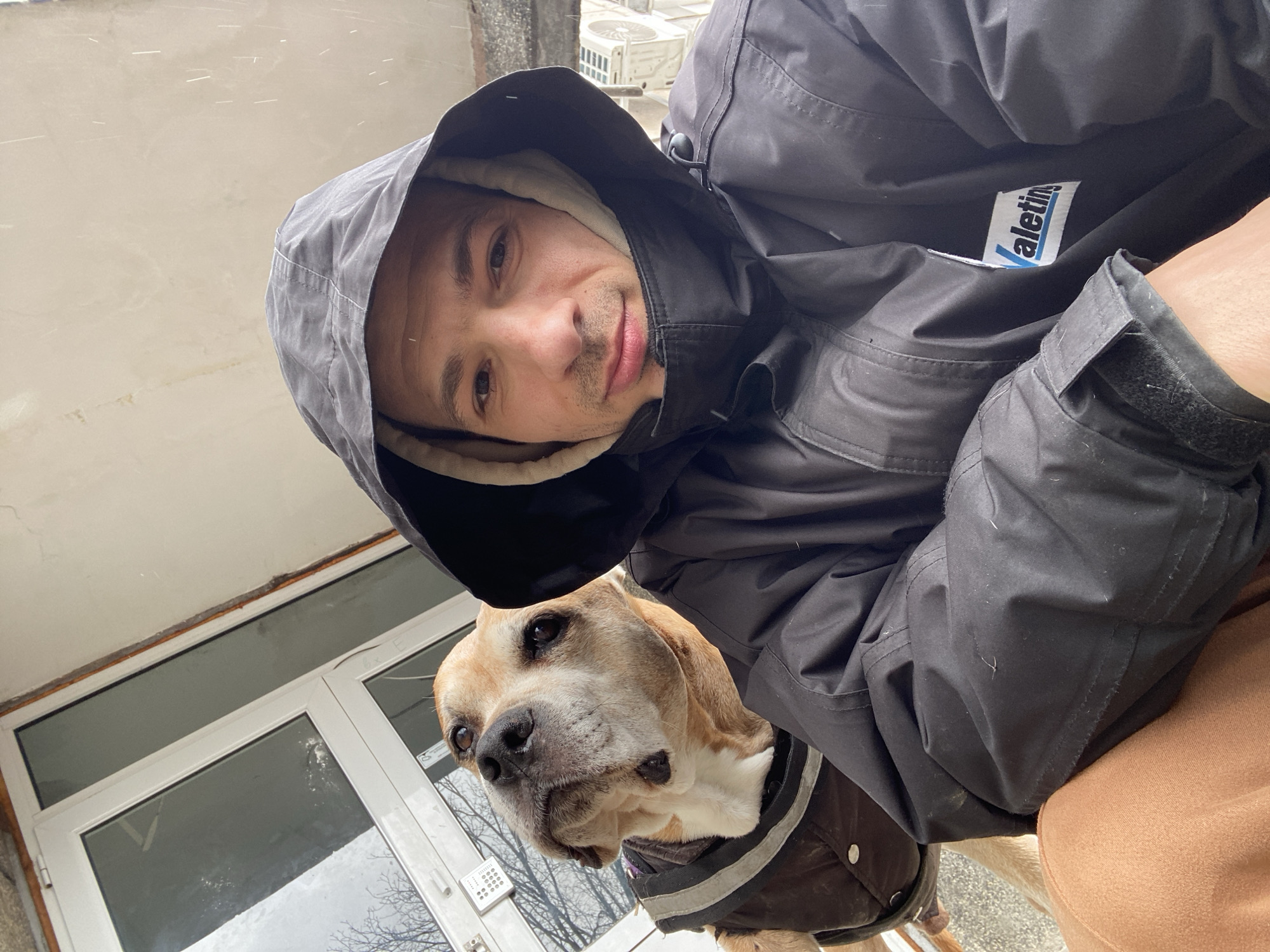 A medium shot captures a man and a dog in an outdoor setting, likely Pleven, Bulgaria. The man, wearing a dark-colored, possibly black, jacket with the word "Valetin" visible on the chest, is looking directly at the camera. He has a hood pulled up over his head, revealing a lighter-colored lining around his face. His expression is neutral. He is holding a tan-and-white dog close to him, the dog wearing a dark-colored vest. The dog looks off to the left of the frame. The background includes a windowed door, partially visible in the lower-left, and a light-colored exterior wall. A couple of HVAC units are partially visible in the background. The lighting indicates an overcast day, and what appear to be light snow flakes are falling.