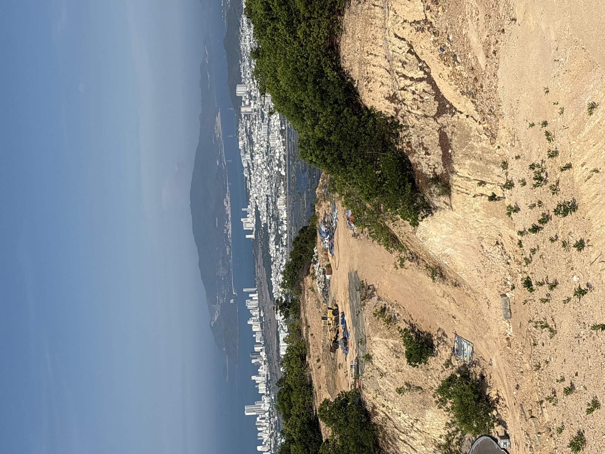 The image shows a steep, arid hillside in Nam Nha Trang Ward, Vietnam, with sparse vegetation and exposed soil. A road or construction site is visible on the hillside, marked by construction vehicles, debris, and building materials. The city of Nha Trang is visible in the background along the coast, with numerous high-rise buildings and the ocean beyond. The sky is clear and blue.