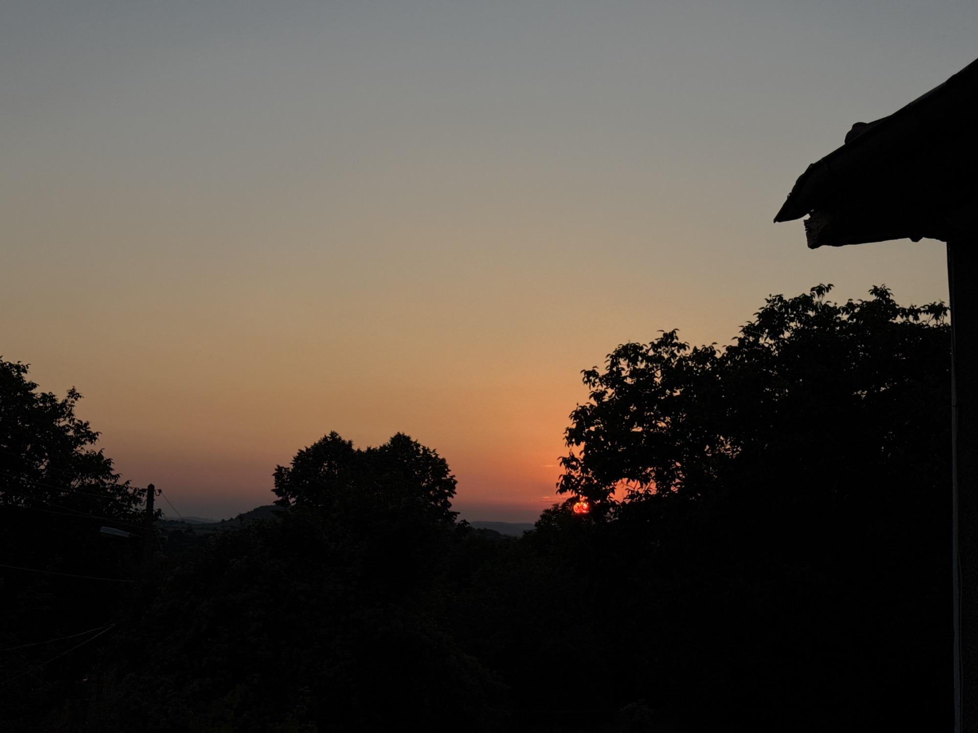 The image captures a silhouetted landscape at dusk, with a gradient sky transitioning from pale blue at the top to warm orange and red hues near the horizon. The setting sun is visible as a bright orange orb peeking through the dark foliage of trees. A portion of a dark-colored roofline is prominent in the upper right corner of the frame, suggesting the presence of a building. The foreground and midground are dominated by the dark silhouettes of trees and vegetation, obscuring much of the terrain and any potential human activity or structures within the city of Krushevo, Bulgaria. The scene is peaceful and devoid of any discernible human presence or action.