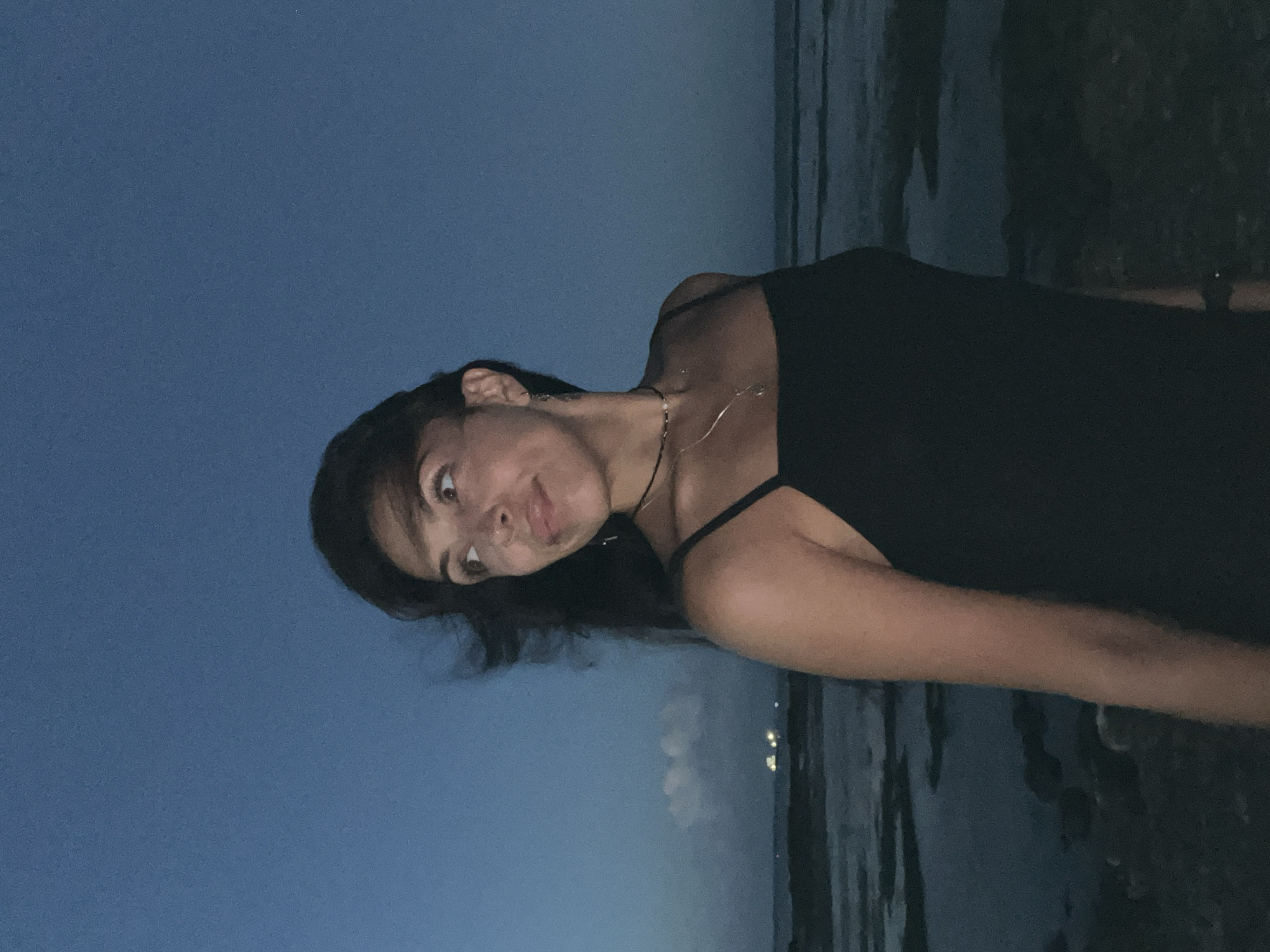 A woman wearing a black tank top stands near the coast at night. She is facing the camera with a slightly tilted head and a playful expression. In the background, the sky is dark blue, transitioning to a darker tone near the horizon. There is a body of water behind her, likely the ocean, with faint lights from what appears to be the city of Kuta Selatan, Indonesia, visible in the distance.