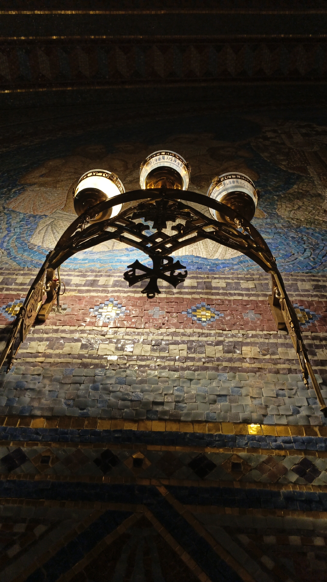 An eye-level shot, taken from the ground looking upward, features a three-light chandelier mounted on a tiled wall. The chandelier is constructed of dark metal and has an arched shape. In the center of the arch is a hanging Chi Rho symbol. The chandelier fixtures hold three glowing globes.
The wall behind the chandelier is covered in small, square tiles arranged in a decorative pattern. The tiles create horizontal stripes of various colors, including blues, reds, and purples. At the top of the wall, the tile pattern transitions into a depiction of human figures.
The photograph was taken in the city of Sevastopol, Ukraine.