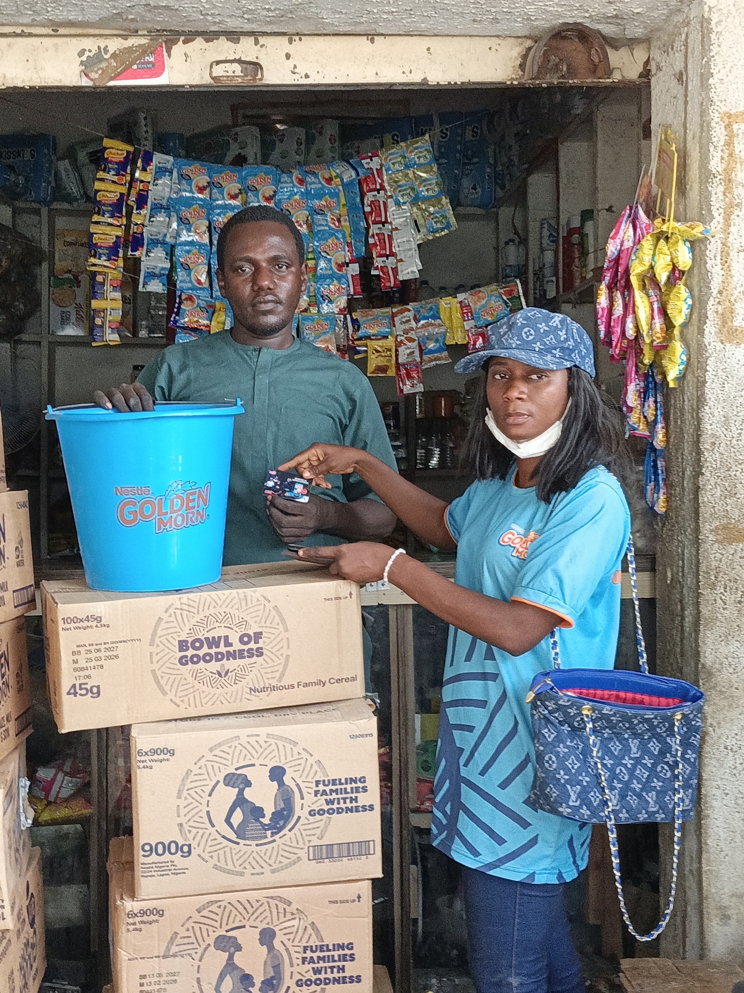 The image captures a scene inside a small retail store in Kawaji, Nigeria. A man stands behind a display of goods, while a woman wearing a blue t-shirt and cap stands in front of him. The man is holding a small item in his hand, and the woman is pointing at it. A light blue bucket with "Nestle GOLDEN MORN" printed on it sits on top of several boxes of "BOWL OF GOODNESS" nutritious family cereal. Shelves behind the man are stocked with various snacks and candies. The woman is also wearing a face mask, and carrying a blue bag with a chain strap.