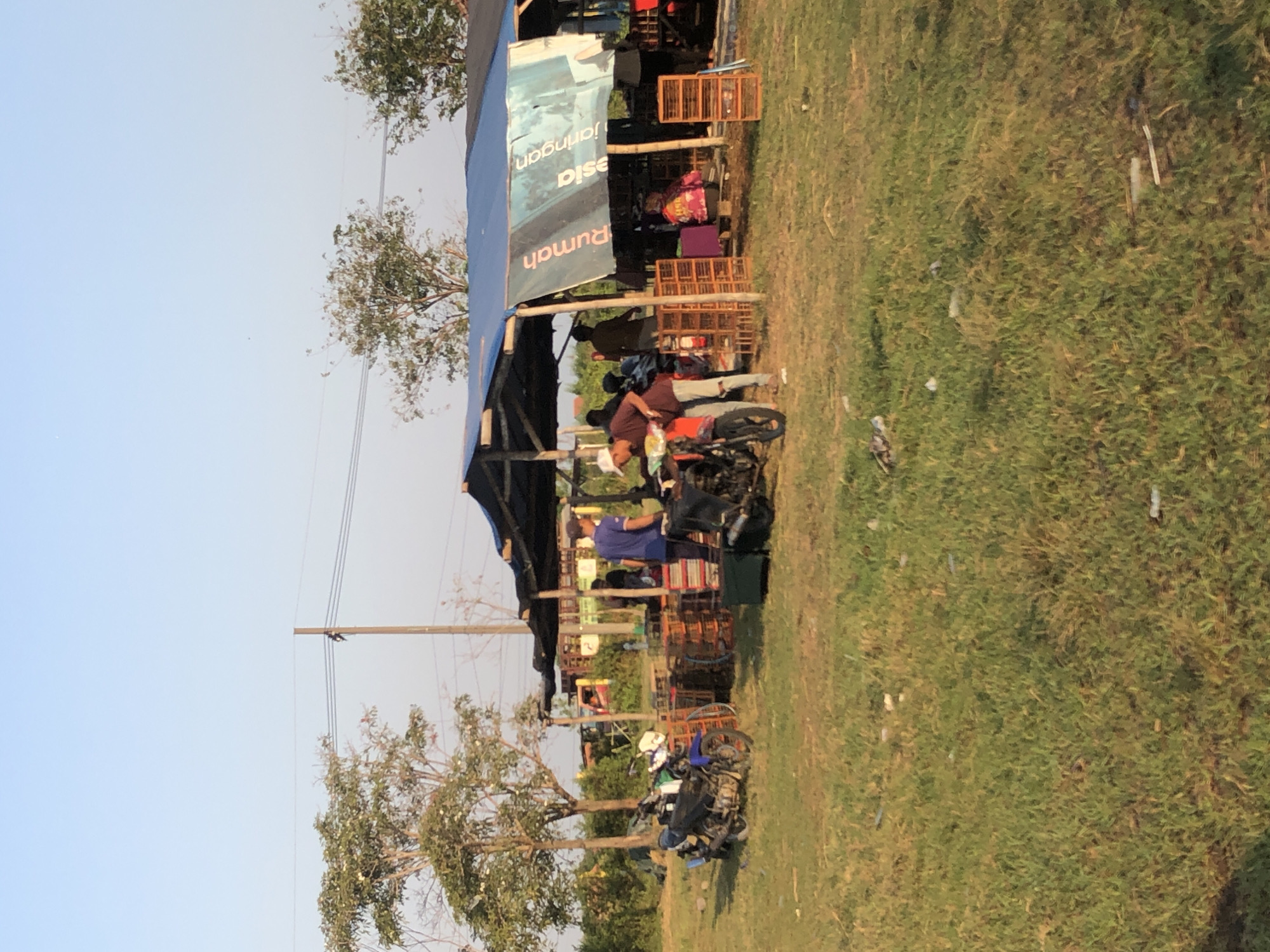 In the city of Gempolsari, Indonesia, an outdoor market scene unfolds under a clear sky. A makeshift structure with a blue tarp roof provides shade for vendors and customers. Several motorcycles are parked on the grassy terrain, one a blue motorcycle on the left, and another darker motorcycle in the center. Wooden crates are stacked in various locations, some near the structure and others around the market area. People are engaged in activities within and around the market stalls. In the foreground, a person wearing a white hat and a maroon shirt is attending to a motorcycle. To the left of the structure, two individuals in blue shirts are visible. Another person, wearing a maroon shirt and a white hat, is standing near a motorcycle. In the background, the silhouettes of more people can be seen, suggesting a bustling market atmosphere. The environment is characterized by grassy fields and scattered trees, indicating a rural or semi-rural setting.
