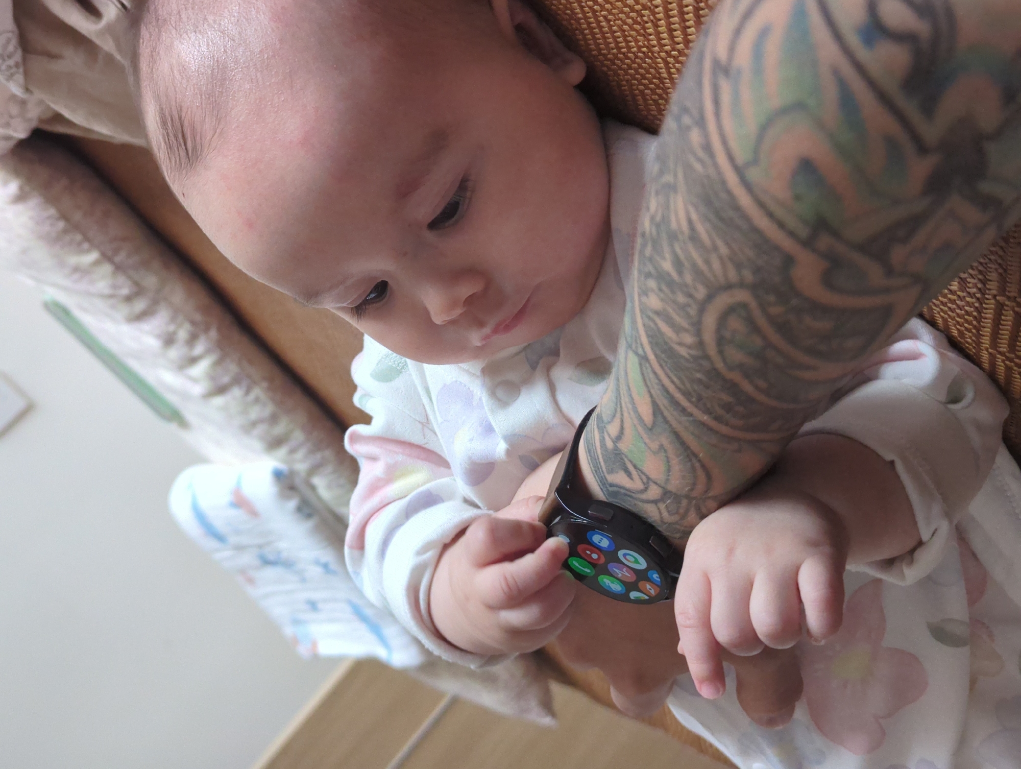 A baby, dressed in a light-colored floral onesie, interacts with a smartwatch on the wrist of a person with extensive arm tattoos. The tattoo extends down the arm, wrapping the forearm. The baby's fingers are touching the watch face. The setting appears to be indoors, with a textured brown surface in the background, and what looks like a patterned pillow to the side. There is no indication of location in Shunde District, China, or any other specific place.