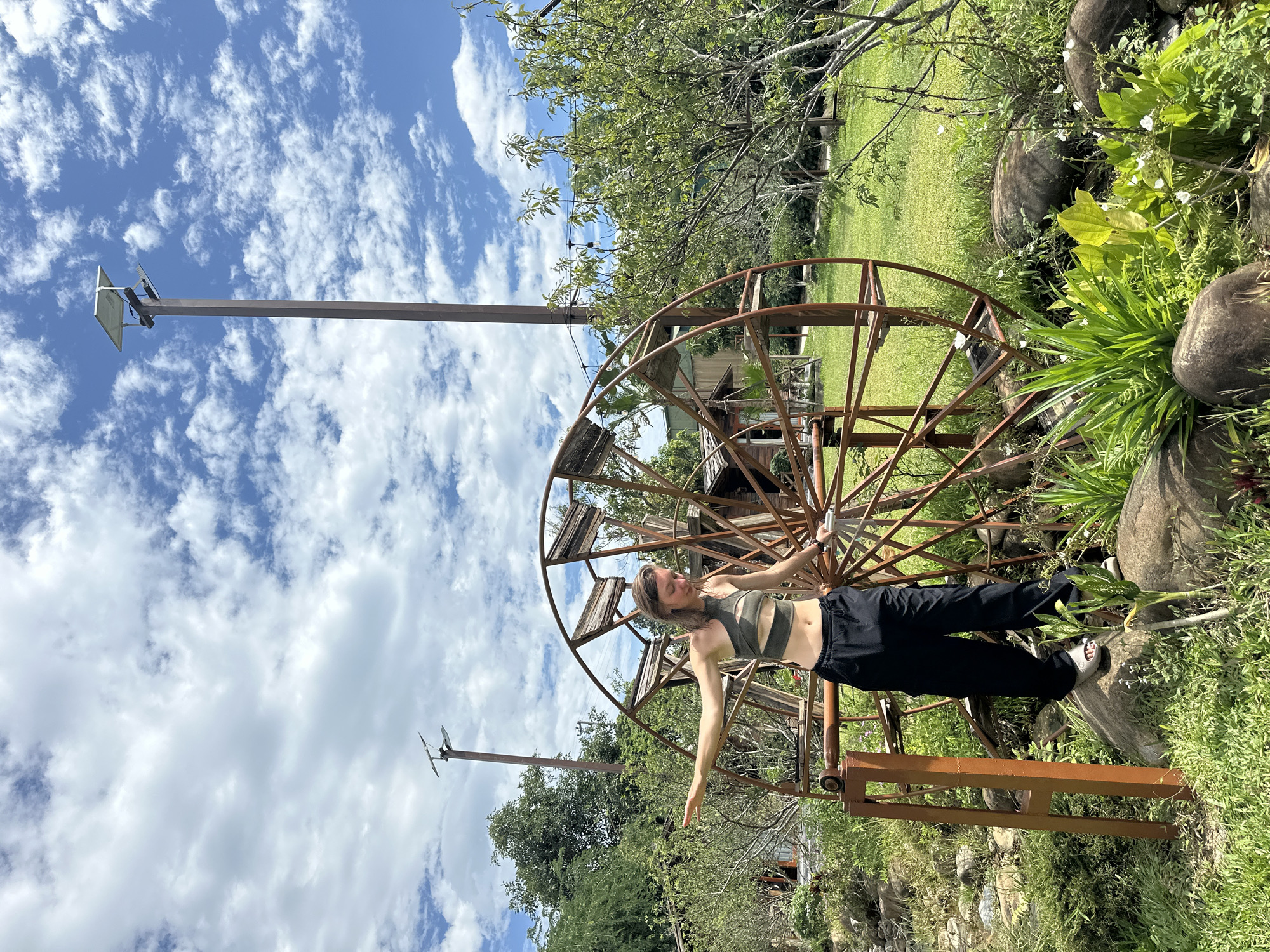 An outdoor scene in Nghia Lo Ward, Vietnam, depicts a young woman balancing near a large, rusty metal decorative water wheel under a bright blue sky with scattered white clouds. The woman, with light skin and brown hair, wears a green one-shoulder crop top with cut-outs and loose black pants, with her arms outstretched for balance. She stands with her right foot on a large rock and her left foot near the wheel's base. The wheel itself is large, made of rusted metal, and features wooden components within its spokes, resting on a rusty metal support structure on the left. The ground around the wheel is covered in lush green grass, various plants, some with small white flowers, and several smooth, dark rocks. In the background, there are numerous trees, some dense foliage, and a rustic wooden building partially visible behind the wheel. Two tall poles equipped with solar panels and light fixtures are prominent against the sky, one on the right and another further in the distance on the left.
