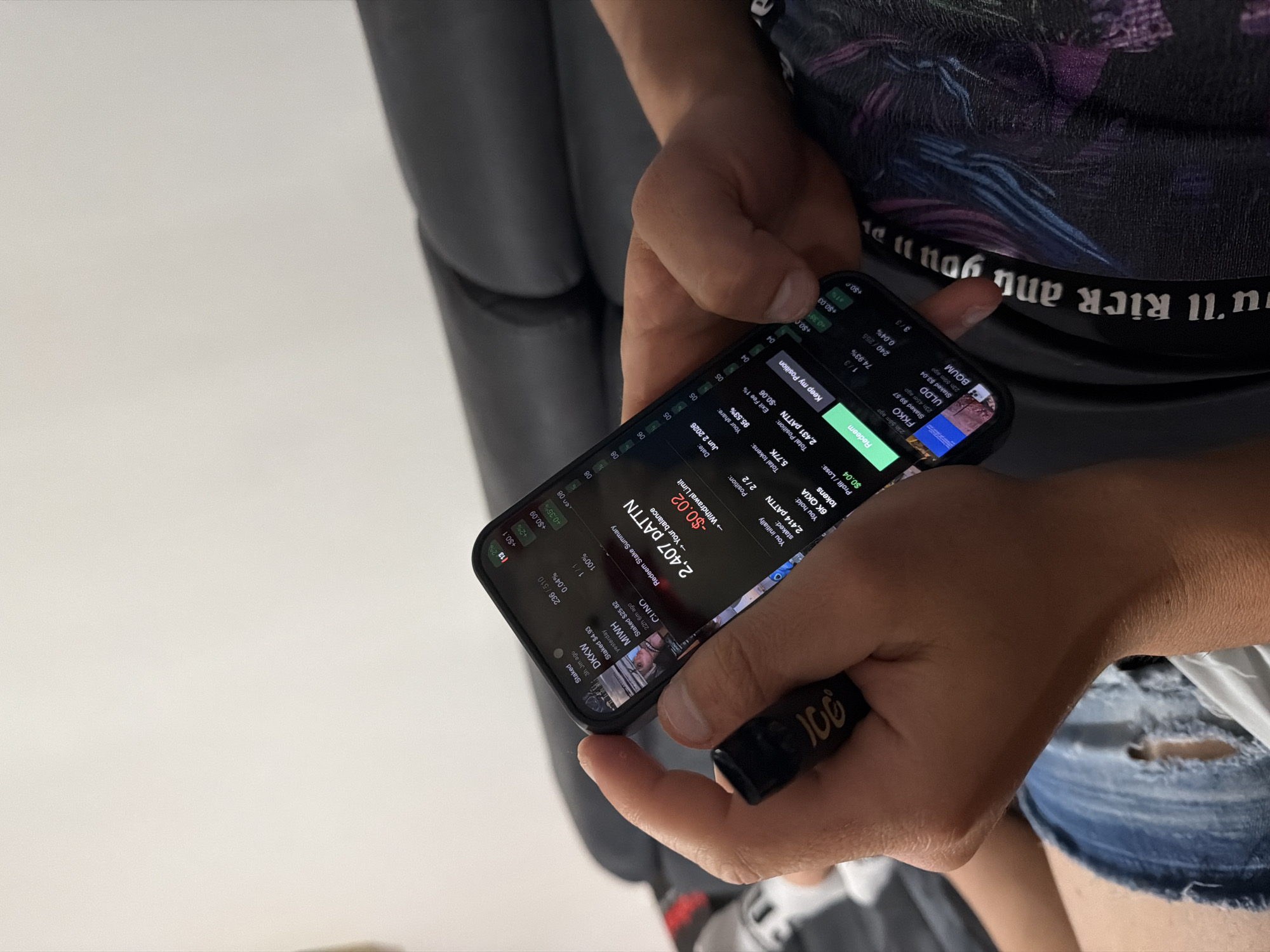 A close-up, eye-level shot captures a person holding and interacting with a smartphone, likely indoors in Plovdiv, Bulgaria. The individual's torso and lower body are partially visible, wearing a dark t-shirt with a colorful graphic design and ripped light blue denim shorts, suggesting a casual setting. Their hands, with visible arm hair, carefully grip a black-cased smartphone, which is tilted to reveal its screen content.
The smartphone displays what appears to be a financial or cryptocurrency staking application. Prominently visible is "2,407 PATTN → Your balance" with a small loss indicated by "-$0.02 → Withdrawal Limit." Other details include "Profit/Loss: $0.04" and various staked assets like "FKKO Staked $9.57," "ULDD Staked $3.04," "CLINO Staked $25.82," "MWH Staked $4.93," and "DKKW," often accompanied by percentages (e.g., "+0.35%", "+0.04%") and time stamps (e.g., "23h 59m ago"). The app also shows "You hold: 6K QKIA tokens" and "Total tokens: 5.77K." Interactive elements include a green "Redeem" button and a "Keep my Position" option, along with "Your share: 95.53%," "Exit Fee 1%," and a "Date: Jun 2 2026."
In the person's right hand, a small black cylindrical object with "100" printed in white is visible. The background consists of a blurred dark grey object, possibly a chair or sofa. Part of the person's t-shirt displays inverted text that reads, "YOU'LL KICK AND YOU'LL." The focus is entirely on the digital interface, with no discernible facial expressions or external environmental cues about the exact time of day or specific location within Plovdiv.