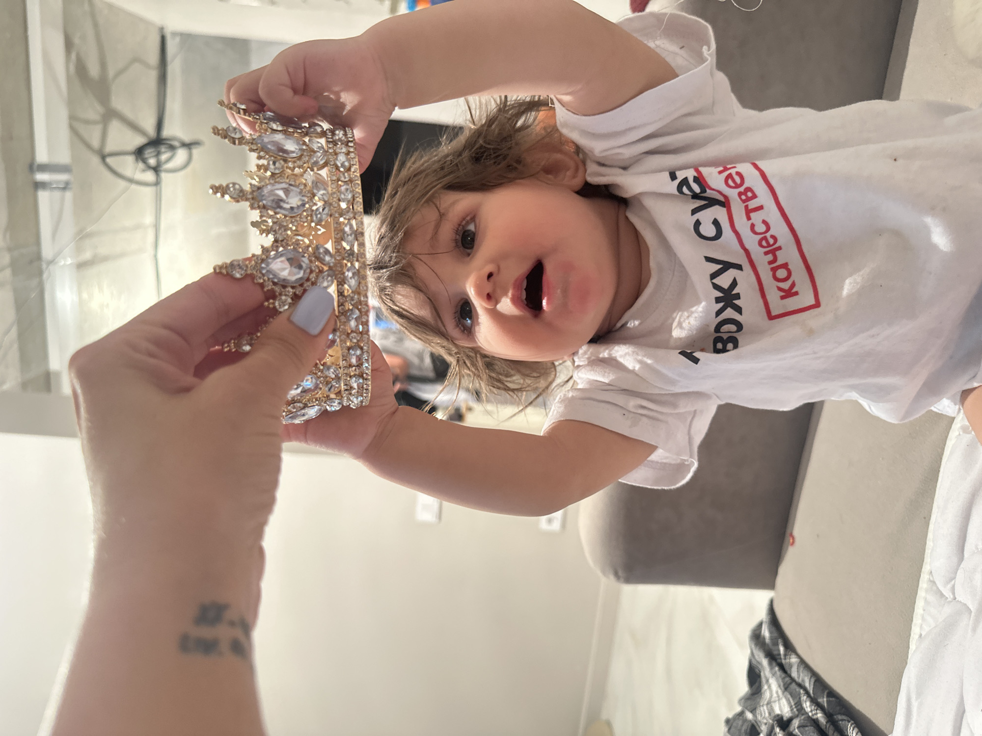 An intimate, slightly rotated, indoor shot captures a playful moment between an adult and a young child, likely in a home setting in Moscow, Russia.
In the foreground, an adult's hand, adorned with light blue nail polish and a blurred tattoo on the wrist, holds a sparkling gold-colored crown. The crown is intricately designed with numerous clear, oval and round jewels that reflect light. The adult is positioned as if presenting or about to place the crown on the child's head.
The child, a baby or toddler with brown hair, looks up towards the crown with a wide-eyed, open-mouthed expression of excitement or wonder. They are wearing a white t-shirt with text printed in Cyrillic. Part of the visible text reads "ВОДЖУ СУ" and within a red rectangular outline, the word "качествен" is clearly visible. This translates to "I drive super high-quality," suggesting a playful slogan on the child's attire.
The background shows a brightly lit indoor environment. A reflection of a ceiling fixture, possibly a fan or light, is visible. There are light-colored walls, and a grey sofa or bed covered partially by a black and white plaid or checked blanket is also present, indicating a cozy, domestic setting. The lighting suggests it is daytime. The overall mood of the image is joyful and tender, capturing a moment of interaction and delight.