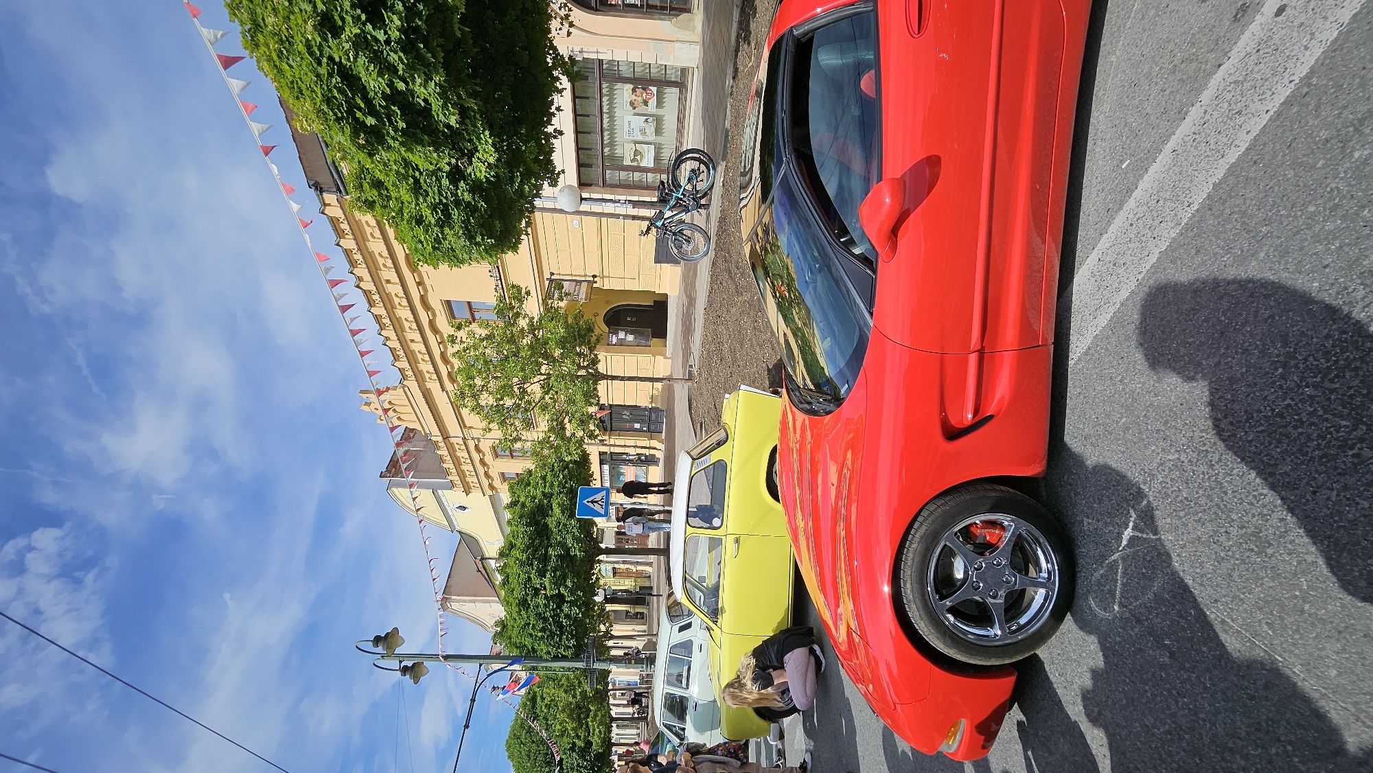This image depicts a sunlit street scene in Prešov, Slovakia, featuring multiple vehicles and pedestrians, suggestive of a public event such as a car show.
In the immediate foreground, the front and right side of a bright red Chevrolet Corvette C5 sports car are prominently displayed, parked on asphalt pavement. Its chrome five-spoke front wheel with red brake calipers is visible. The car has a black targa roof panel. Distinct, elongated shadows of multiple individuals are cast on the asphalt to the lower left and right of the car, indicating strong sunlight from the right side of the frame.
In the midground, to the left of the red Corvette, a bright yellow two-door classic car, consistent with a Trabant 601, is parked. A woman with blonde hair, wearing a black top and light-colored pants, is crouched next to the front left wheel of the yellow car, appearing to observe or interact with it. Further back on the left, another light-colored (possibly light green or blue) classic sedan, resembling a Škoda 100 or similar Eastern European model, is also parked.
The street is lined with trees featuring green foliage, and a blue pedestrian crossing sign with a white 'A' symbol is visible on a pole in the mid-left. Several pedestrians are scattered on the sidewalk and street in the mid-background. On the right side of the street, against a building, a black and turquoise mountain bike is parked next to a lamppost.
The background consists of a row of traditional European-style buildings. The most prominent building, in the center-right, is a multi-story structure painted a warm yellow/ochre color, featuring decorative elements such as crenellated parapet features and geometric patterns below the roofline. Other buildings are visible in various shades of yellow and cream. Red and white triangular bunting is strung across the street between the buildings, indicating a festive or celebratory atmosphere. The sky above is clear blue with sparse white, wispy clouds. Overhead utility wires traverse the upper left corner of the frame.