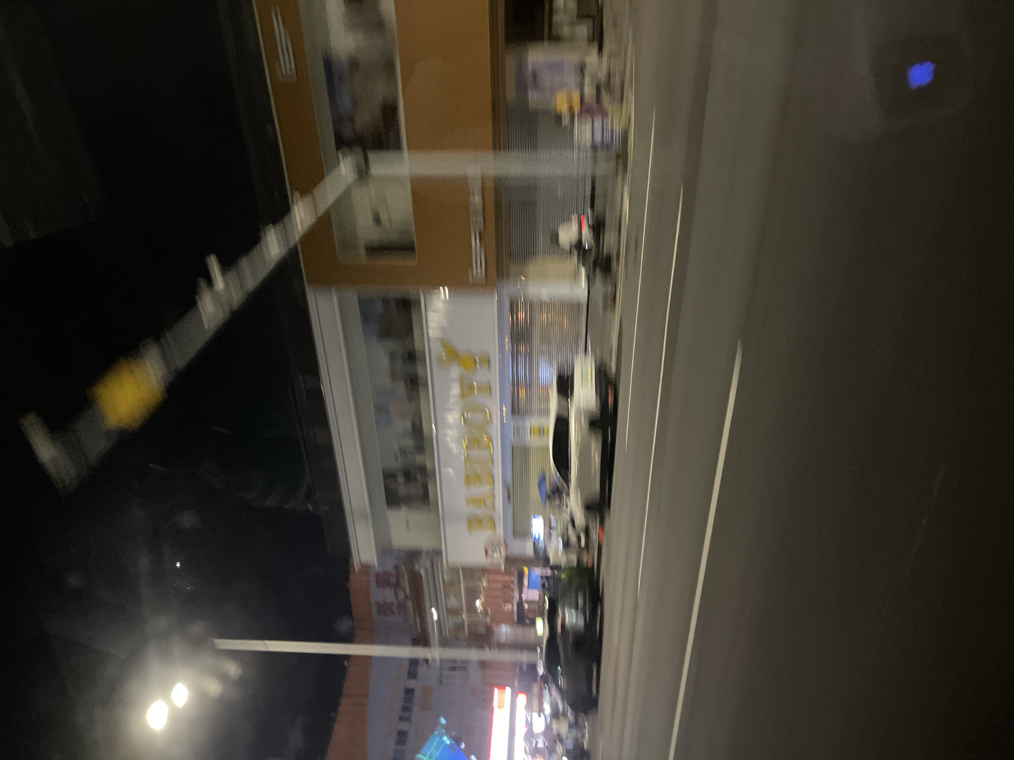 The image captures a street scene at night in Panyu District, China. A building with a prominent sign reading "BABIBOY'S" is visible, featuring yellow lettering. Adjacent to this building, another structure with an orange facade and windows is present. Several vehicles are on the road, including a dark-colored SUV on the left and a white sedan on the right, both appearing blurred due to motion. Streetlights illuminate the scene, casting light on the road and sidewalks. Some individuals are discernible in the background near the buildings, though their features are indistinct due to the low light and motion. The overall impression is of a commercial area at night, with some activity on the street.