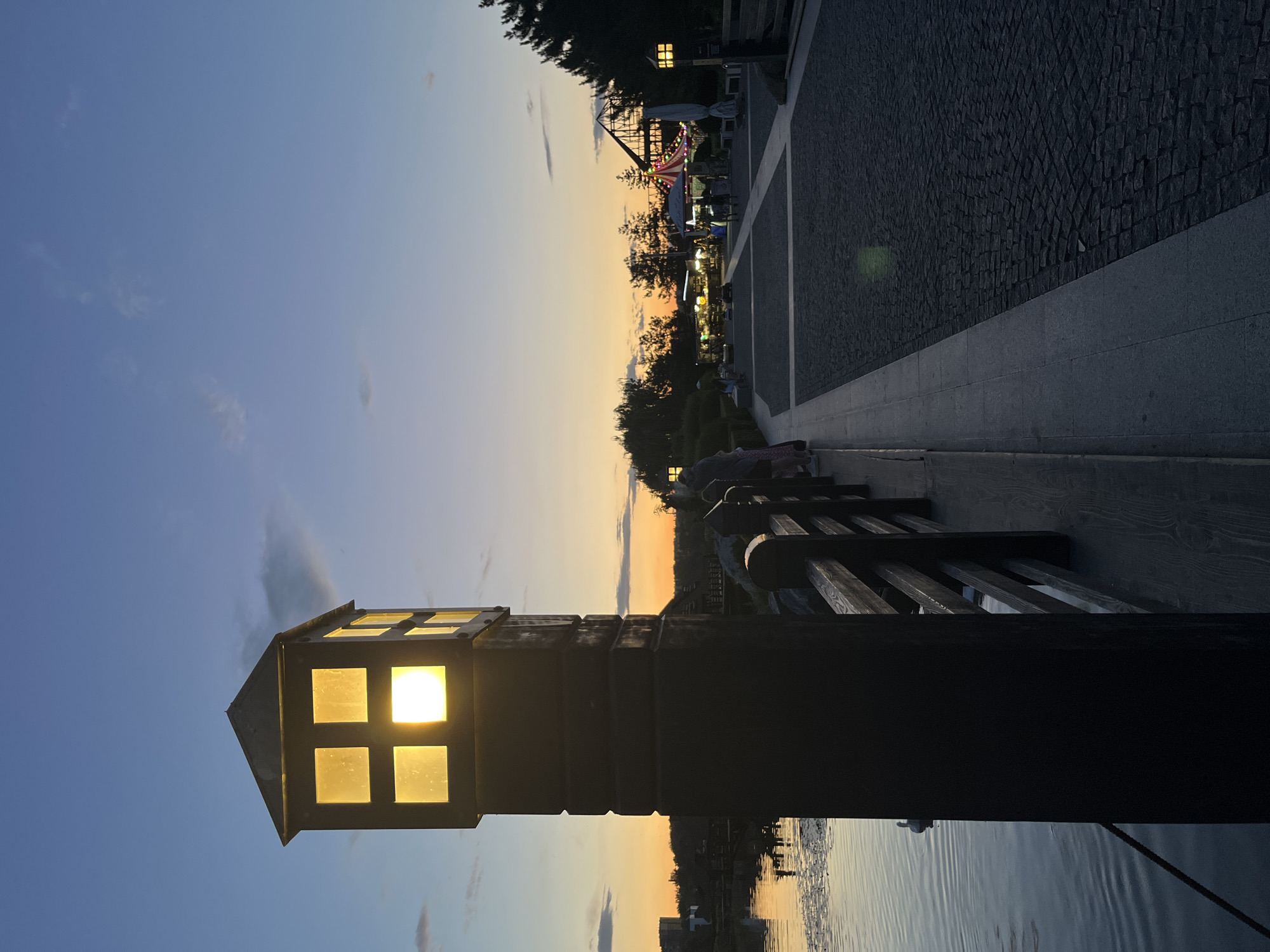 The image depicts a sunset scene at a park-like area with a body of water in Dnipro, Ukraine. In the foreground, a dark, square lamppost with four illuminated glass panes stands to the left, casting a warm glow. To its right, a wooden railing runs along a paved walkway made of cobblestones. The water on the left reflects the twilight sky and the surrounding structures. In the midground and background, several people are visible walking along the paved area, some near tents and stalls that suggest a recreational or entertainment venue. Trees dot the landscape, silhouetted against the colorful sunset sky. The overall atmosphere is serene and inviting, characteristic of an evening in a public park.