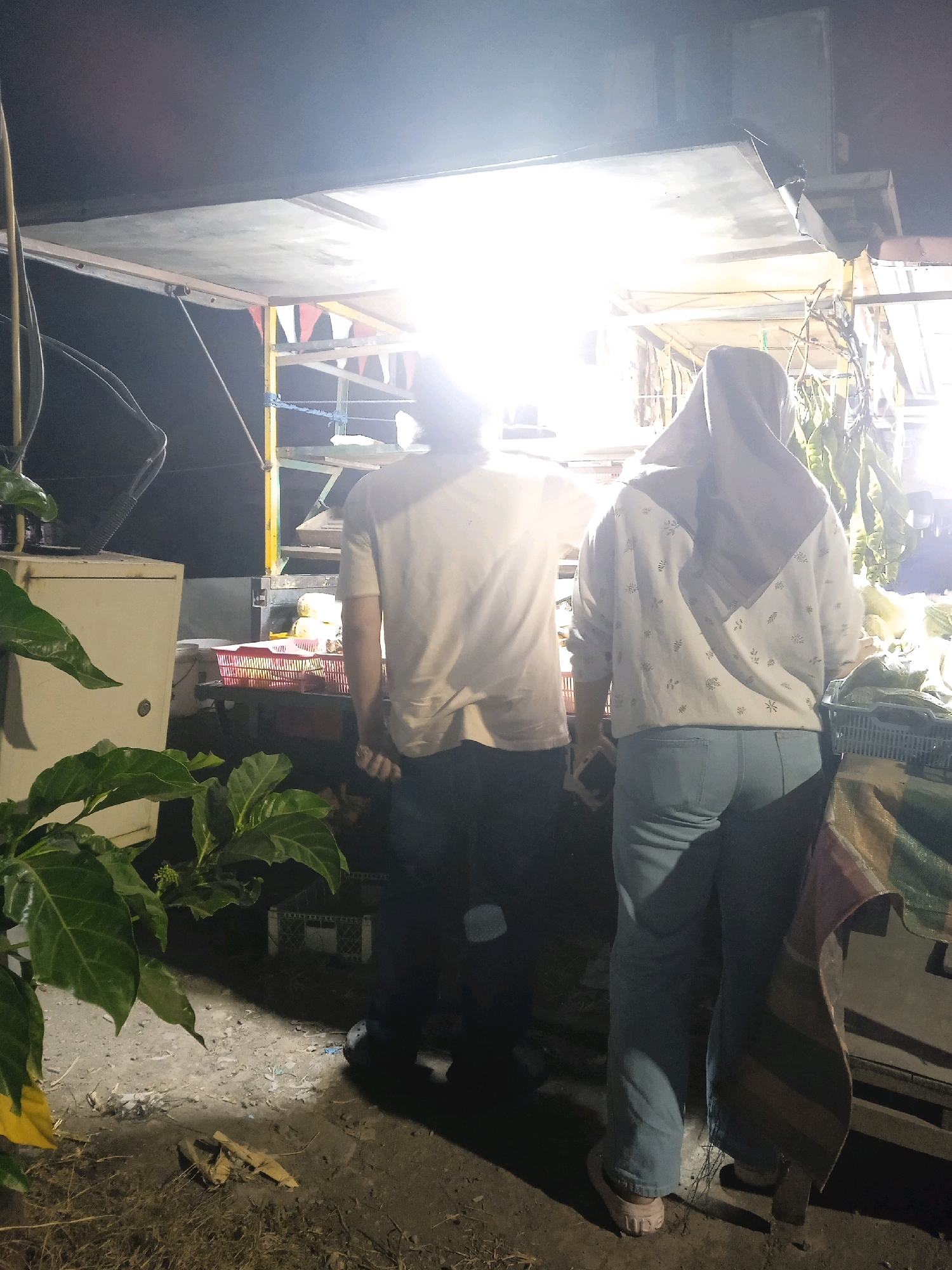 In Langensari, Indonesia, at night, two individuals stand at a food stall illuminated by bright overhead lights. The stall displays produce, including what appears to be corn. A male figure, facing away from the camera, is wearing a light-colored t-shirt and dark pants. Beside him, a female figure, also facing away, is wearing a headscarf and a patterned sweater over light blue jeans. The ground is dirt and littered with debris, including dried leaves and a discarded piece of cardboard. Green foliage from a plant is visible in the foreground.