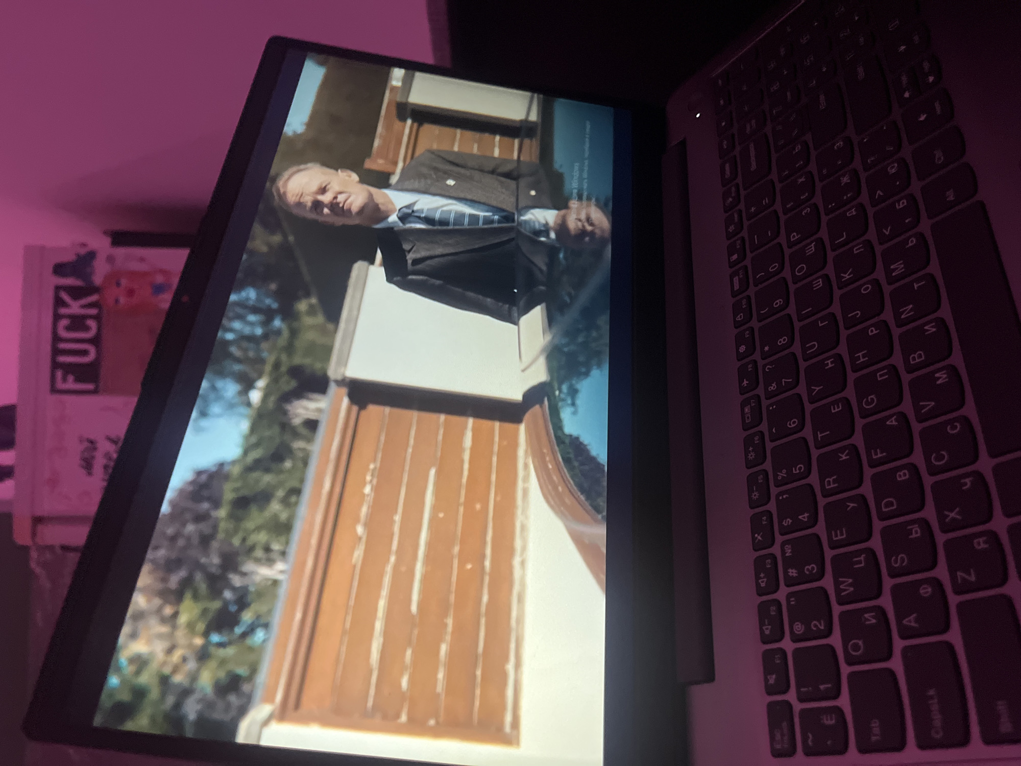 A laptop screen displays a scene featuring a man in a suit and tie standing in front of a building with brown paneling, possibly set outdoors with trees visible in the background. A sticker with the word "FUCK" in large letters is partially visible behind the laptop screen. The laptop keyboard shows Russian characters. The lighting in the room appears dim, with a pinkish hue, suggesting an indoor setting. The geographical location of the scene on the screen in the city of Saint Petersburg, Russia.