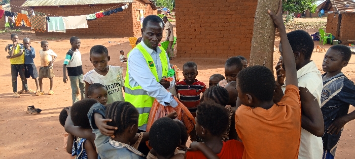 An outdoor scene in Ngalula, Democratic Republic of the Congo, depicts an adult male distributing items to a large group of children on a reddish-brown dirt ground. The male, with dark skin and short dark hair, wears a light blue long-sleeved collared shirt beneath a bright yellow-green high-visibility vest with green insignia. He is positioned centrally, bending forward slightly, holding an orange translucent plastic bag in his left hand and extending a small, green and orange object with his right hand towards a dense cluster of approximately 15-20 children.
The children, ranging in age from toddlers to pre-teens, are gathered tightly around the man, with many looking directly at the items he is distributing or reaching out. Their attire varies, including t-shirts, shorts, and dresses of different colors. In the background to the left, several other children stand at a distance, observing the activity. A clothesline strung between two single-story, unpainted brick structures displays assorted garments. Additional unpainted brick buildings, some with corrugated metal roofs, are visible throughout the background, indicating a village or residential area. A small tree trunk stands upright near the central group. The ground is unpaved, consisting of dry, reddish-brown earth. Scattered objects, including a pair of dark shoes and distant green plastic chairs, are present on the ground. The scene is illuminated by natural daylight.
