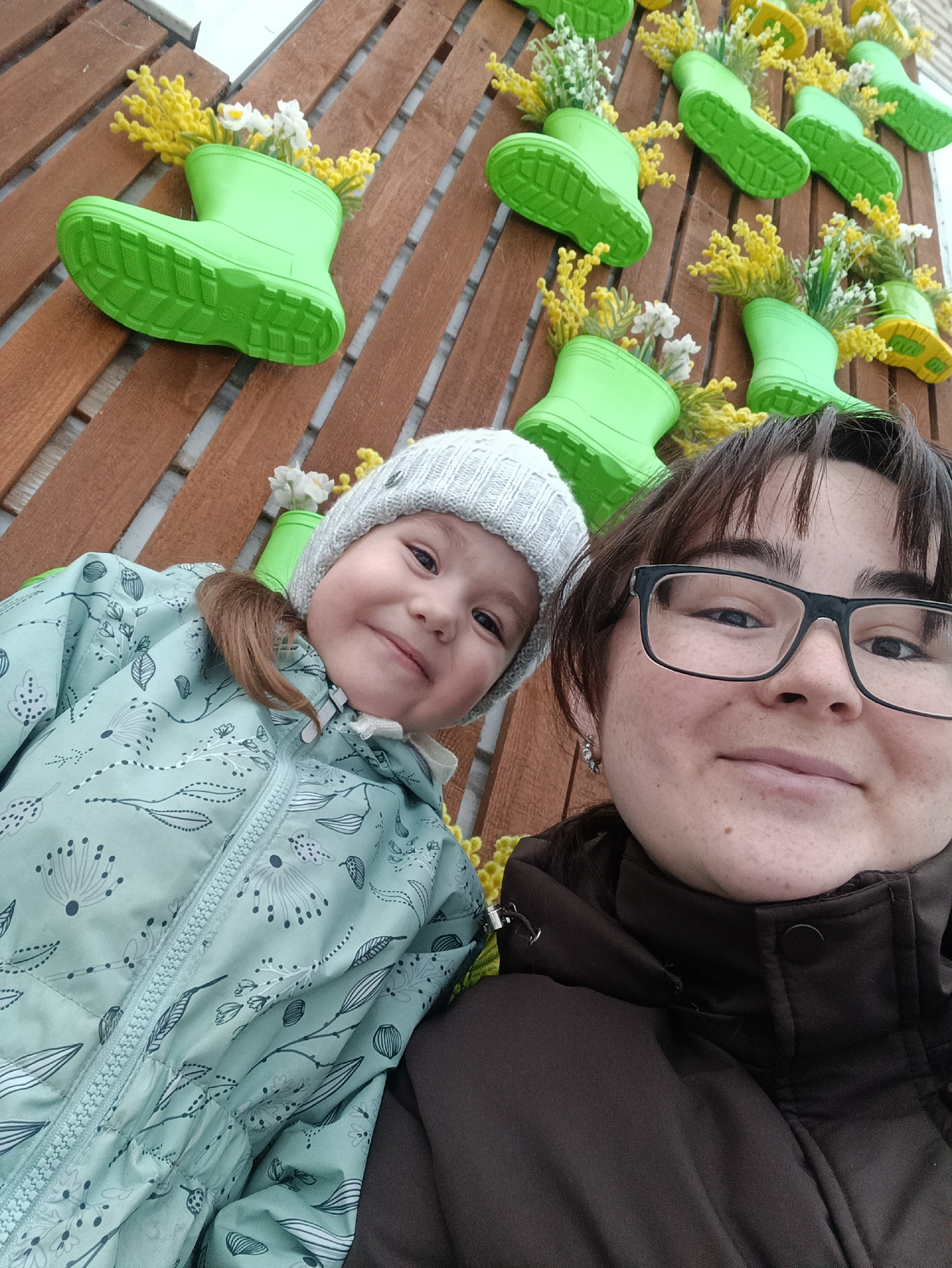This image, captured in Chaikovsky, Russia, presents an upward-angled close-up of two individuals against a decorative wall. The subject on the right is an adult female with dark brown hair and black-framed glasses, wearing a dark brown jacket. Freckles are visible on her face, and she has a silver earring in her left ear. Adjacent to her on the left is a young child, wearing a light grey knitted beanie with small studs and a light teal jacket adorned with a white and grey floral/leaf pattern. The child's light brown hair is partially visible in a small ponytail. Both subjects are looking directly towards the camera, exhibiting smiles. The background comprises a vertical wooden slat wall, stained reddish-brown, to which numerous bright green small-sized rubber boots are affixed. Each boot contains artificial yellow mimosa-like flowers and small white flowers. The boots are oriented variably, with some displaying yellow sole elements.