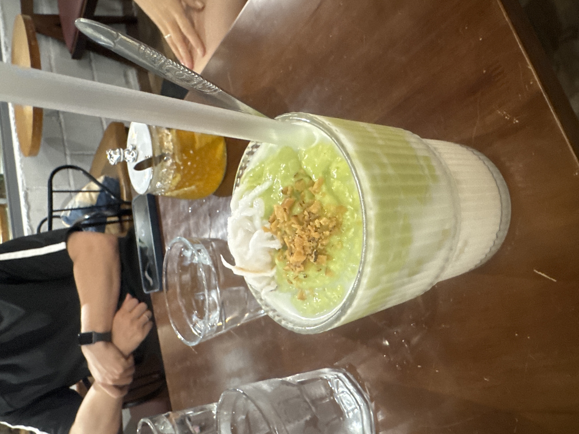 In Bac Nha Trang Ward, Vietnam, a close-up shot captures a layered beverage in a ribbed glass, topped with shredded coconut and chopped nuts. The drink, with a green smoothie on top of a white, milky layer, is served with a white straw and a spoon. Nearby, a glass jar filled with honey and a small silver lid with a cat figurine sits on a brown wooden table. Two other glasses of water are visible, along with hands resting on the table. In the background, a person wearing a black t-shirt with white stripes is seated, their arms crossed, and a smartwatch on their left wrist. The environment suggests a casual dining setting.