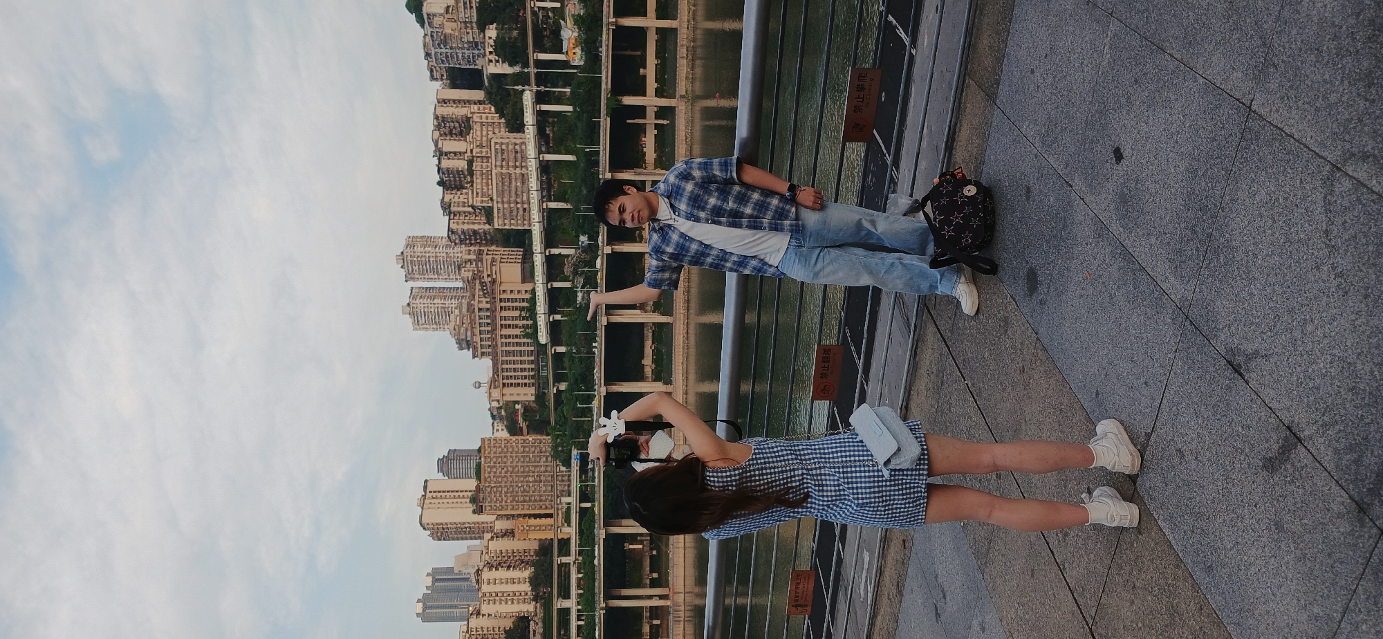 A medium shot depicts a man and a woman standing on a gray tile surface with a metal railing, with a backdrop of buildings and a body of water.
The woman, seen from the back, is wearing a blue and white checkered dress with white sneakers and has long, dark hair. She holds a camera with white accessories near her face.
The man is wearing a blue and white checkered shirt, a white t-shirt, light blue jeans, and white sneakers. He has a backpack near his feet and extends his right arm.
In the background, there is a river or body of water with a bridge or structure running across it. Buildings of varying heights and designs are visible, suggesting a city location. The sky is visible with the sun going down. Based on the architectural style, and geographical features, the location is likely Yuzhong District in China.