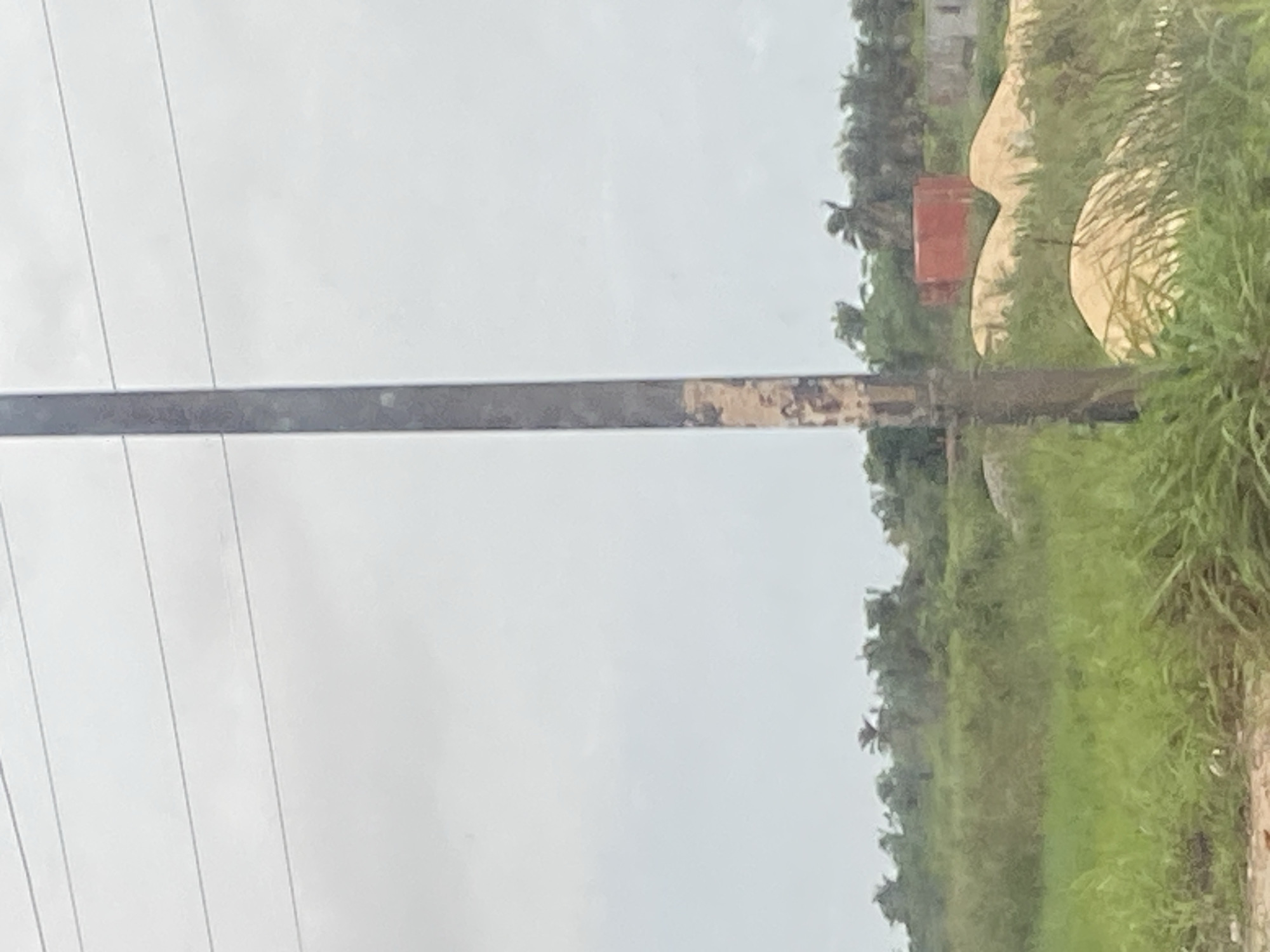 The image depicts a power line pole situated in a landscape that appears to be on the outskirts of Lagos, Nigeria. The pole, vertical and centrally positioned, shows signs of wear with faded markings on its surface. In the background, lush green vegetation, characteristic of the Nigerian climate, dominates the scene. Beyond the greenery, several mounds of sand are visible, suggesting construction or land development activities. A prominent red container stands amongst the sand piles, further indicating ongoing work. The sky is overcast, contributing to the generally muted tones of the environment. The scene is devoid of any people, and the overall impression is one of an undeveloped or developing area within the urban sprawl of Lagos.