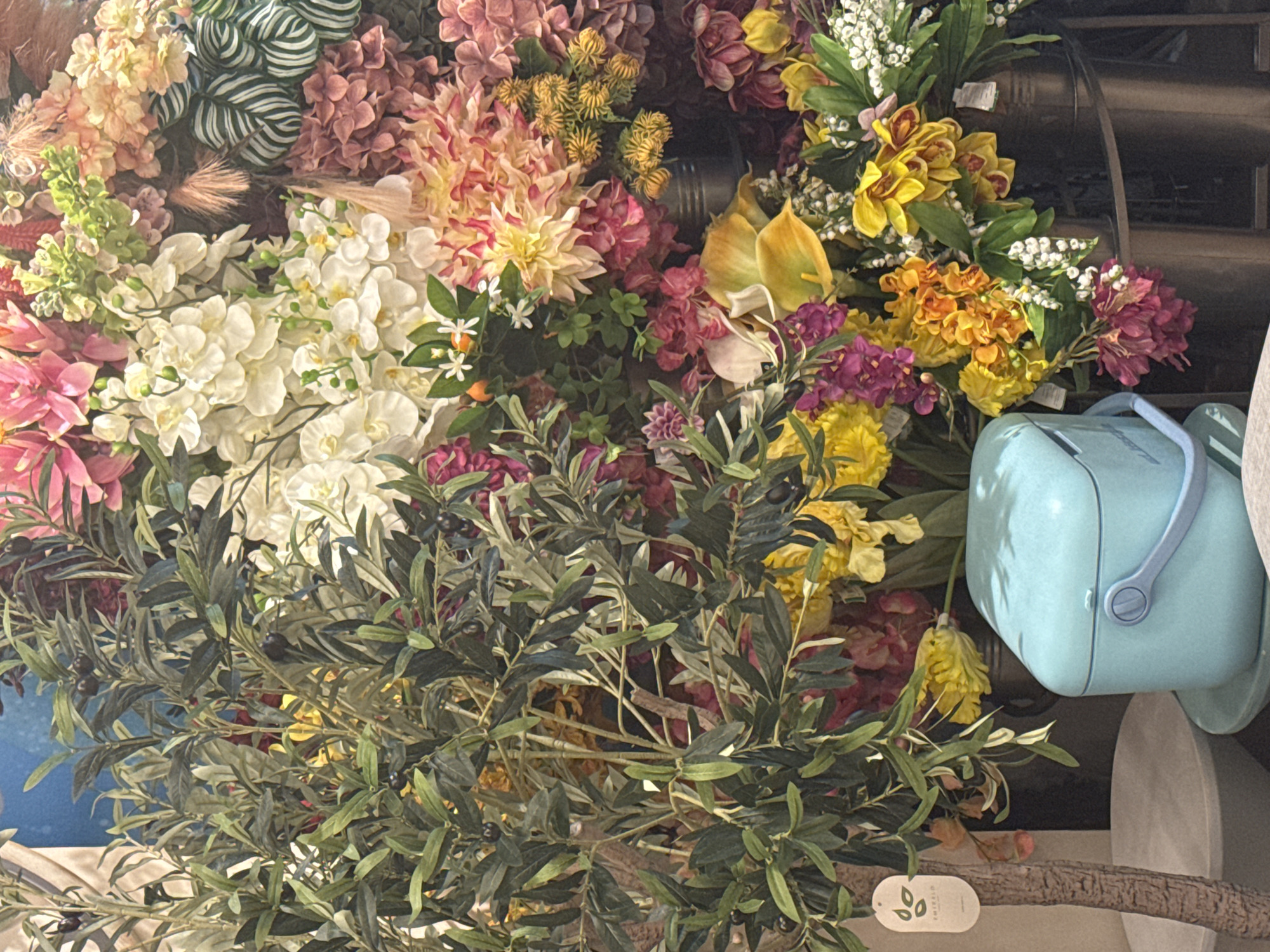 A vertical, eye-level, medium shot captures a vibrant display of artificial flowers, predominantly in shades of pink, yellow, orange, white, and purple, with lush green foliage. On the left side of the frame, an artificial olive tree with dark green leaves and small black olives is positioned in front of the floral arrangement. The tree has a textured brown trunk and a small white tag with green lettering is attached to one of its branches. In the foreground, towards the bottom center, a light blue plastic cooler with a light blue handle is visible. The cooler sits on a light grey circular object, which in turn rests on a dark grey surface. The background consists of a dense arrangement of various artificial flowers and greenery, creating a colorful and textured backdrop. The lighting appears to be even, with no harsh shadows, suggesting an indoor setting, possibly a retail display. The image is sharp and well-focused, highlighting the intricate details of the artificial flora.