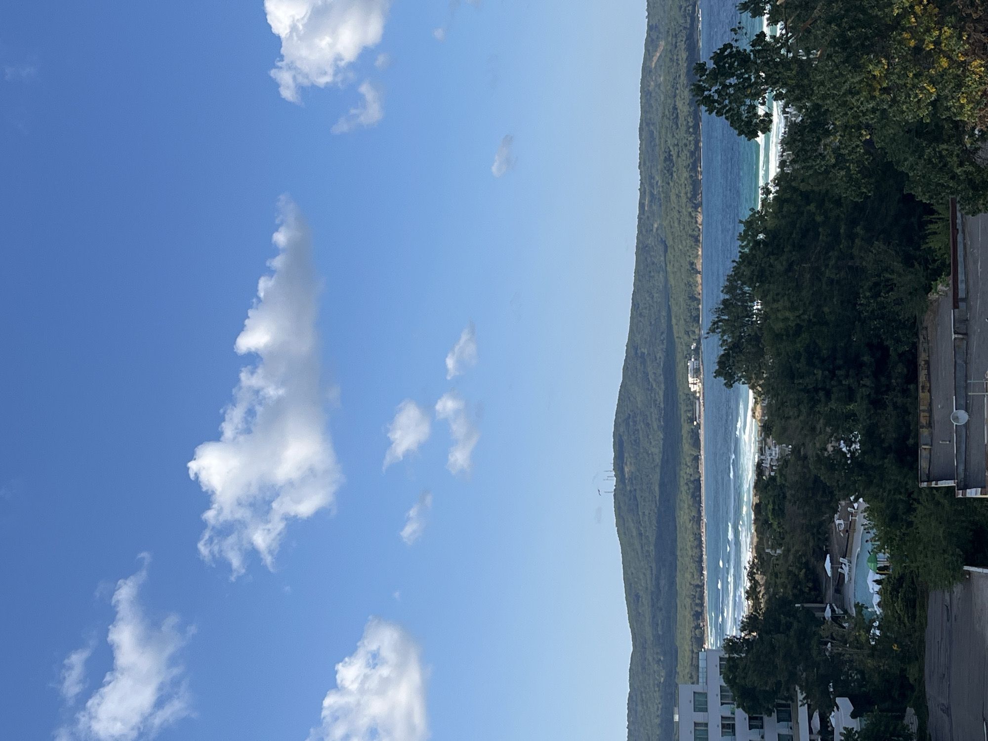 The image depicts a scenic view of the coast in Primorsko, Bulgaria. In the foreground, rooftops of buildings are visible, one with a satellite dish. Lush green trees dominate the mid-ground, partially obscuring a resort area with a swimming pool and lounge chairs. Further back, a sandy beach with people and umbrellas stretches along the coastline, met by the bright blue waters of the sea. Waves are breaking on the shore. In the distance, a verdant, tree-covered hill rises, topped with communication towers. The sky above is a clear blue, dotted with fluffy white clouds.