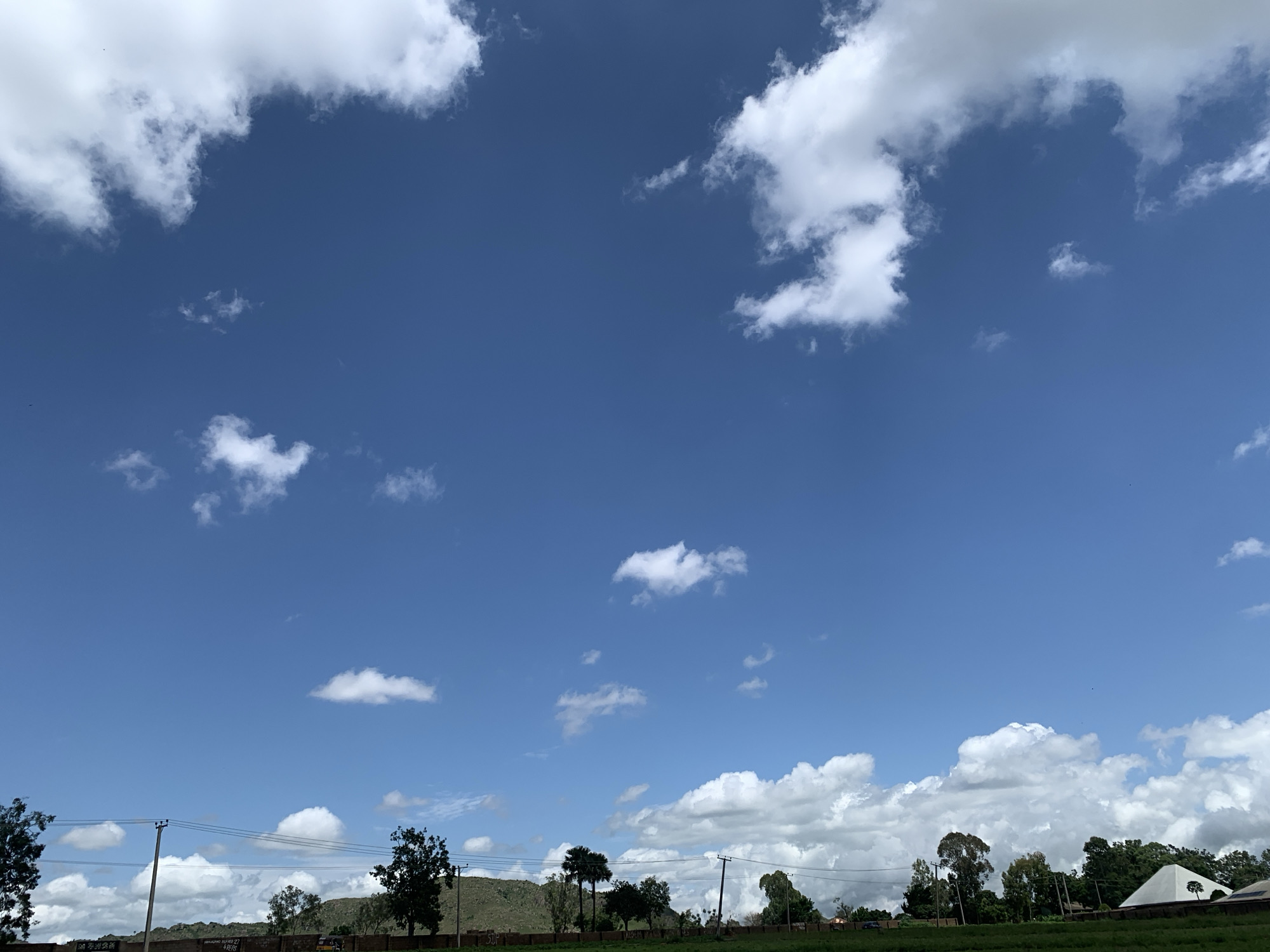 The image captures a bright, clear blue sky with scattered white clouds above a rural landscape. In the foreground, a low wall runs horizontally across the bottom of the frame, partially obscured by a grassy area. Several utility poles with wires extend upwards from behind the wall. Trees, varying in size and density, dot the landscape, some appearing to be located in the mid-ground and others further back. A gently sloping hill or mound is visible in the distance, covered in greenery. The scene suggests an outdoor environment, possibly on the outskirts of Kuru Karama, Nigeria, given the presence of utility infrastructure and the natural, undeveloped terrain. No people or specific actions are depicted in the image.
