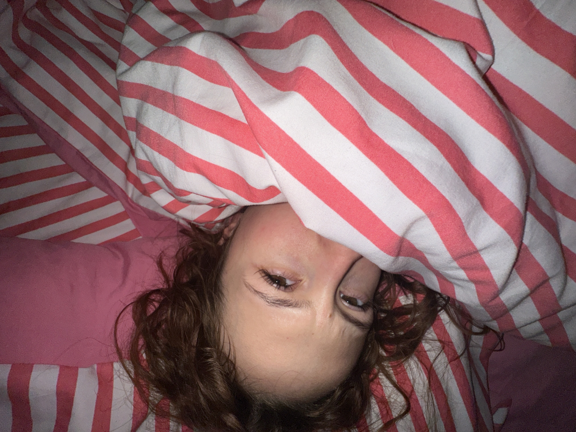 A person is lying on a bed covered with striped pink and white sheets. The person's face is partially covered by the sheets. The person has brown hair. The photograph does not contain any information about location in Novi Sad, Serbia.