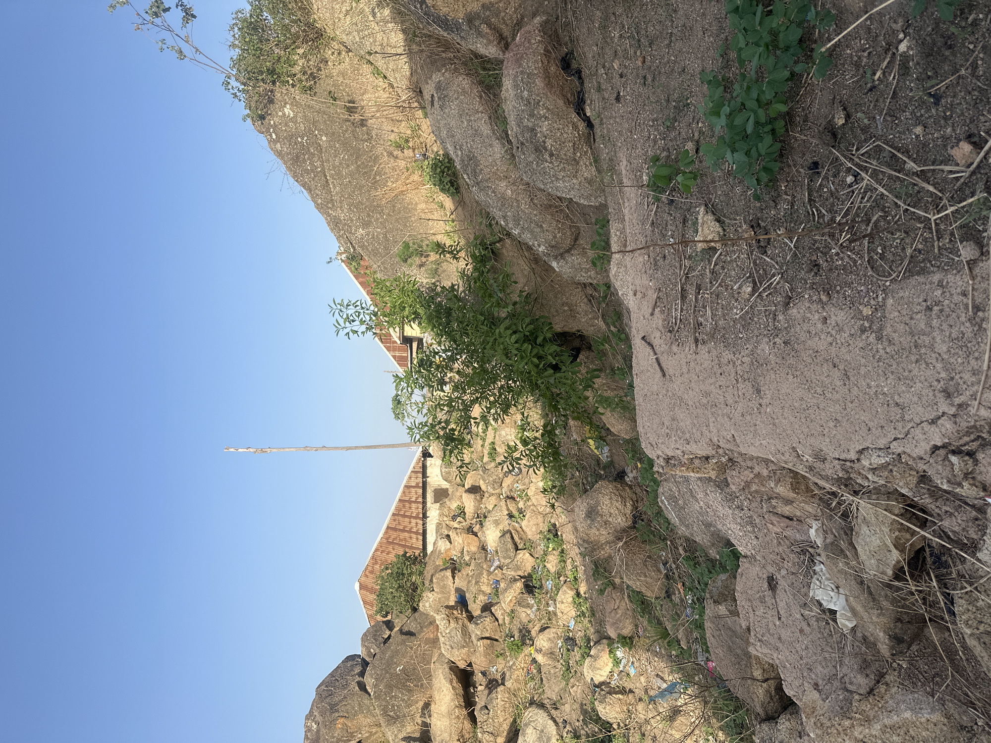 The image depicts a rocky landscape in Jos, Nigeria. The foreground consists of large, weathered rocks and boulders, interspersed with sparse vegetation, including small trees and shrubs. Litter and debris are scattered among the rocks. In the midground, a building with a corrugated metal roof is partially visible behind the rocks. A tall, slender pole or stick extends upwards into the clear blue sky. The image is taken from a low angle, emphasizing the height and scale of the rocks.