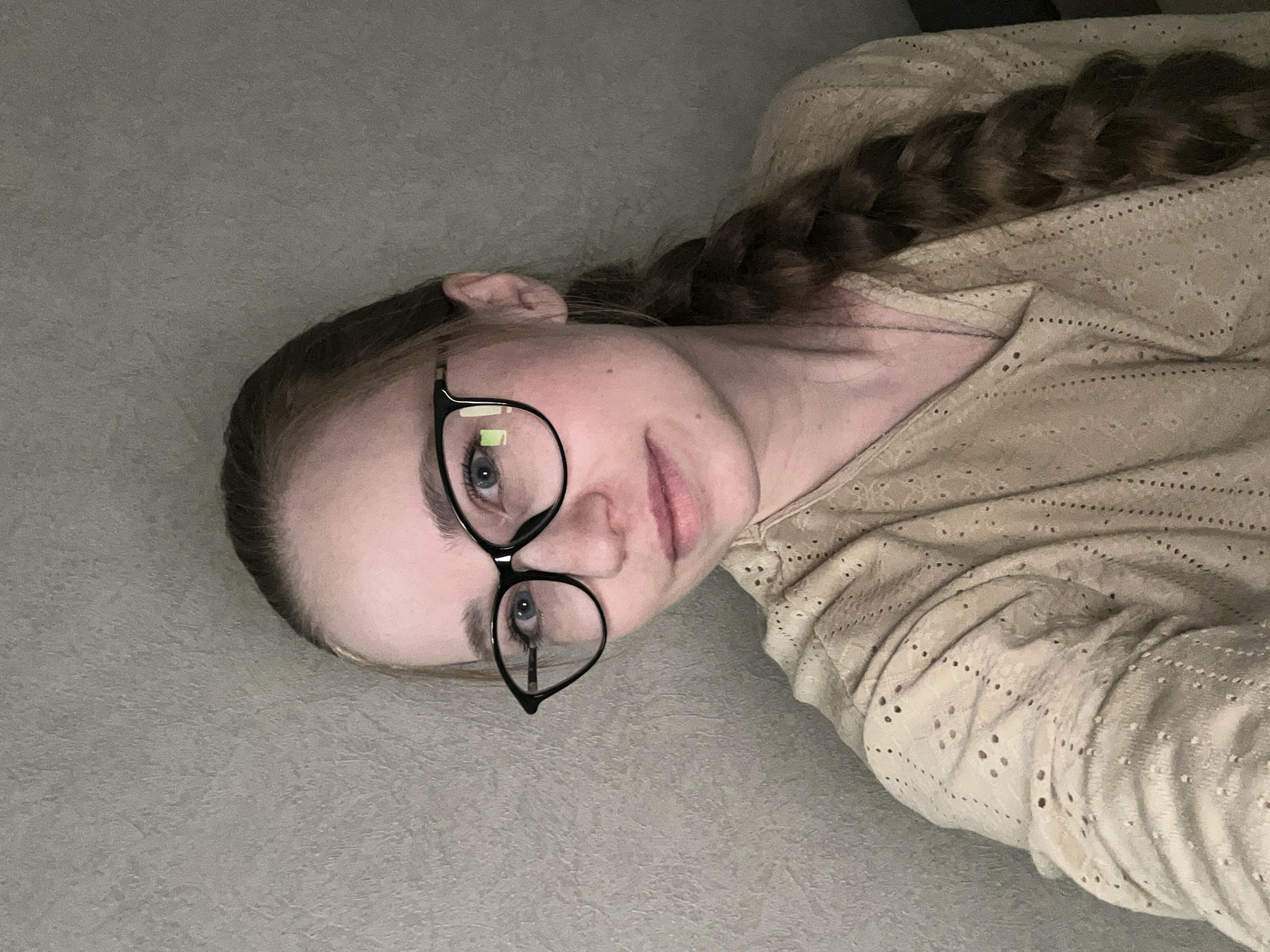 A young woman is lying on a gray surface, possibly a bed or floor, with her head turned towards the viewer. She is wearing black-rimmed glasses. Her hair is light brown and braided, with the braid extending behind her. She is wearing a light beige or cream-colored shirt with patterned perforations. The background is plain gray, likely a wall or flat surface. There is no indication of the city of Saint Petersburg, Russia, or any specific location.