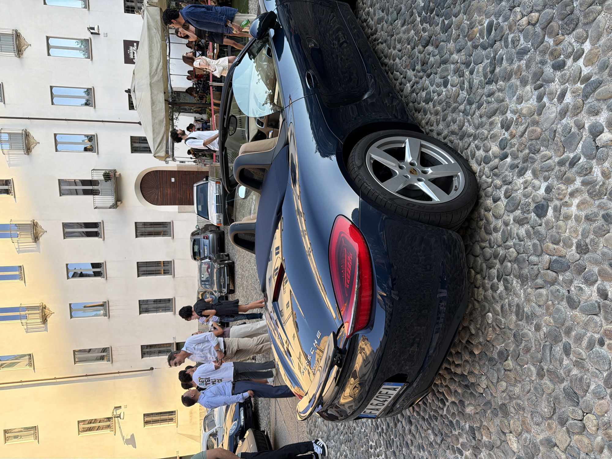 In an outdoor, cobblestone-paved urban setting, a dark blue Porsche Boxster convertible is parked, facing right. The car's rear spoiler and taillights are visible. The license plate reads "FA 640FA". In the background, several people are gathered, conversing and standing near buildings. Some are near a covered outdoor seating area with umbrellas. The buildings have multiple windows and balconies. Other vehicles, including a white Mercedes-Benz, are also present. The lighting suggests it is late afternoon or early evening.