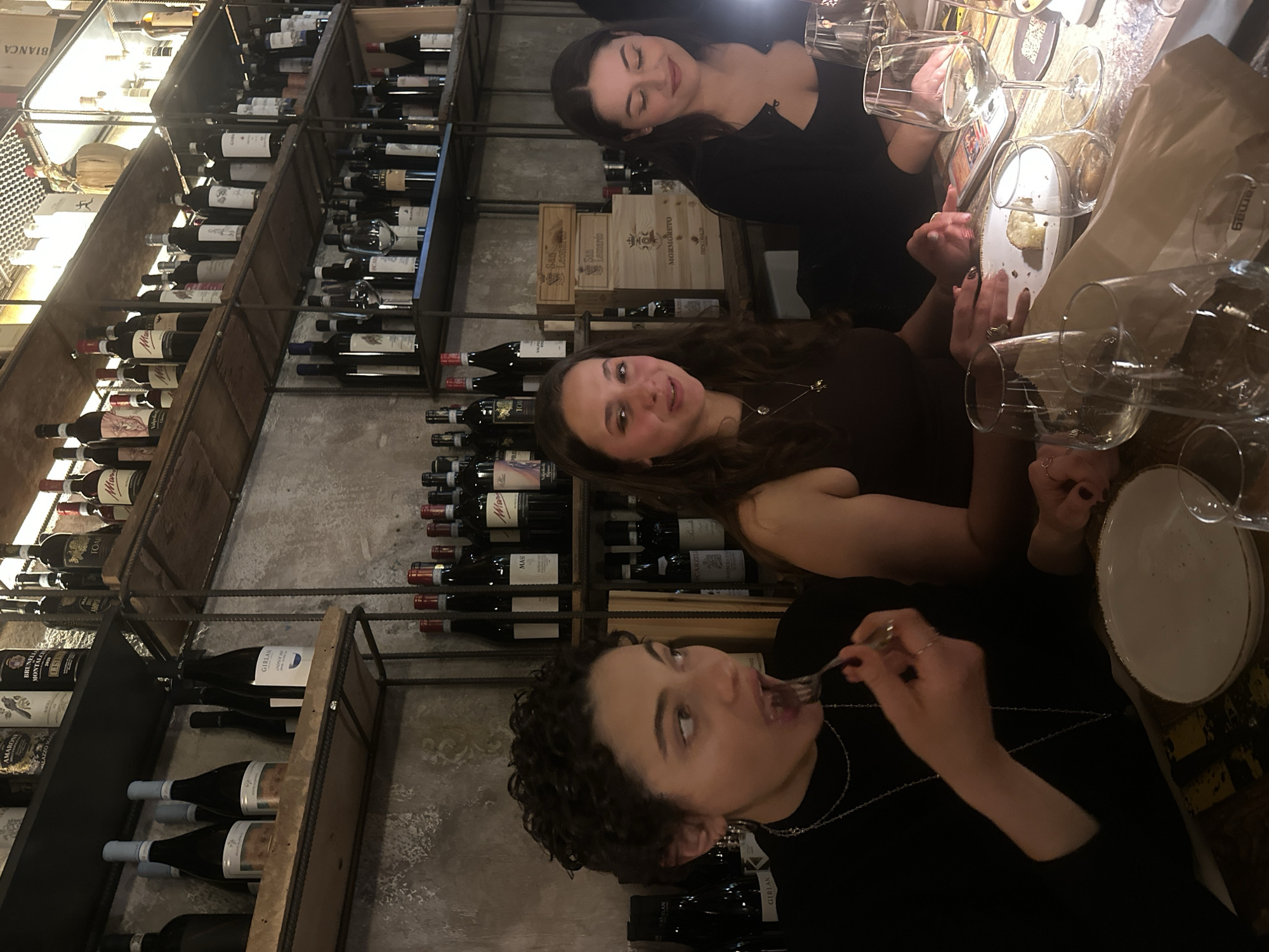 Three young adult women are seated at a distressed wooden table in a dimly lit Italian restaurant or wine bar, characterized by concrete-like textured walls and extensive industrial-style shelving displaying numerous bottles of Italian wine. The establishment is illuminated by warm, ambient lighting.
From left to right:
1. A woman with short, dark, curly hair, wearing a black long-sleeved top and multiple silver chain necklaces, is actively consuming food. She holds a fork, laden with a dark food item, elevated towards her open mouth. She wears a ring on her right ring finger.
2. A woman with long, dark brown wavy hair, dressed in a dark brown sleeveless top with a gold-colored pendant, is seated, looking forward with a slight smile and her mouth partially open. Her hands rest on the table near a white plate containing a small piece of bread or food. She wears a ring on her right ring finger.
3. A woman with long, dark brown straight hair, clad in a black top, is looking downwards towards the table with a subtle smile. Her face is distinctly illuminated from the right by an off-camera light source, likely a mobile phone screen. She wears a ring on her left ring finger.
The table is set with multiple empty and partially filled wine glasses of various designs, white ceramic plates with gold rims, and cutlery. One wine glass holds a small quantity of white wine. A folded paper napkin is also present. The background features floor-to-ceiling metal and wood shelving systems densely packed with a variety of wine bottles, including identifiable Italian brands such as "Girlan," "Masi," "Tommasi," "Scarzello," "Maramis," "San Leonardo," "Brunello di Montalcino," and "Mormoreto." Several wooden wine crates are also stacked on shelves.
Given the prominent display of numerous Italian wine labels and the general aesthetic consistent with an Italian enoteca or restaurant, the scene is likely situated in such an establishment within Milan, Italy, as specified. However, no specific signage or unique Milanese architectural elements are visible to forensically pinpoint an exact street address or district within the city.